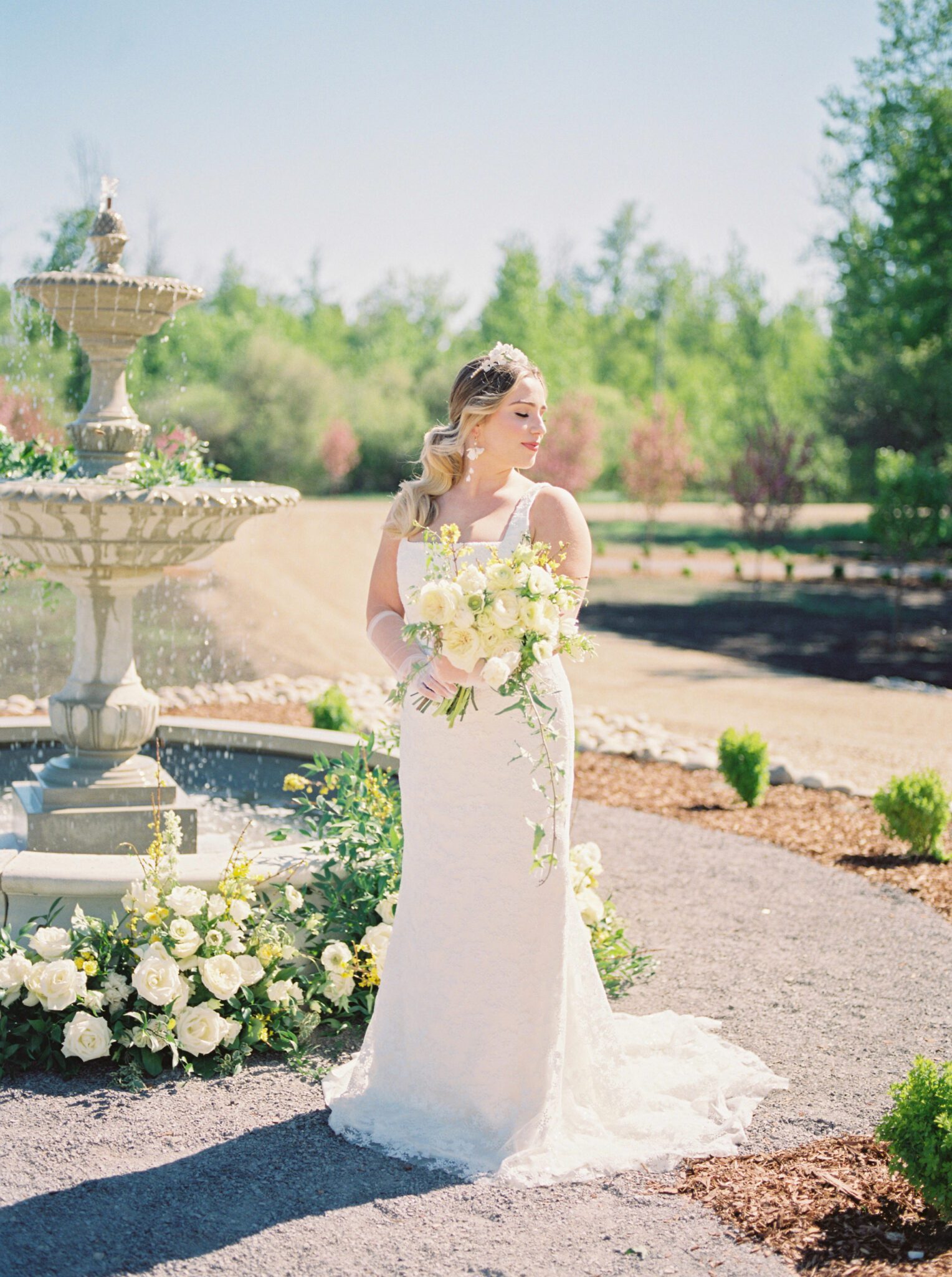 Bride standing in front of the water fountain at Sparrow Lane Events,  holding pale yellow, ivory and white floral bouquet of roses and peonies for the perfect spring wedding inspiration. 
