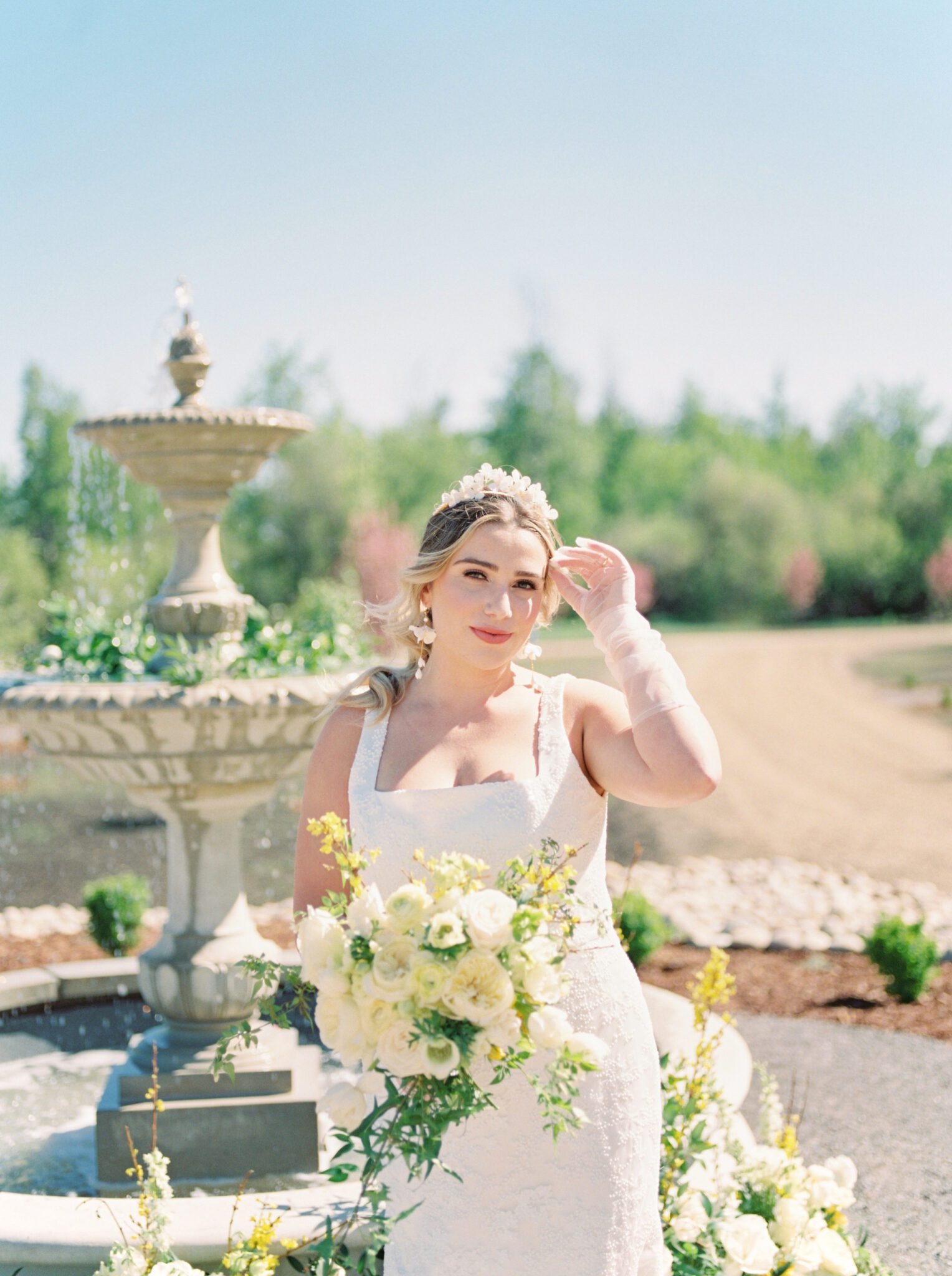 Bride standing in front of the water fountain at Sparrow Lane Events,  holding pale yellow, ivory and white floral bouquet of roses and peonies for the perfect spring wedding inspiration. 
