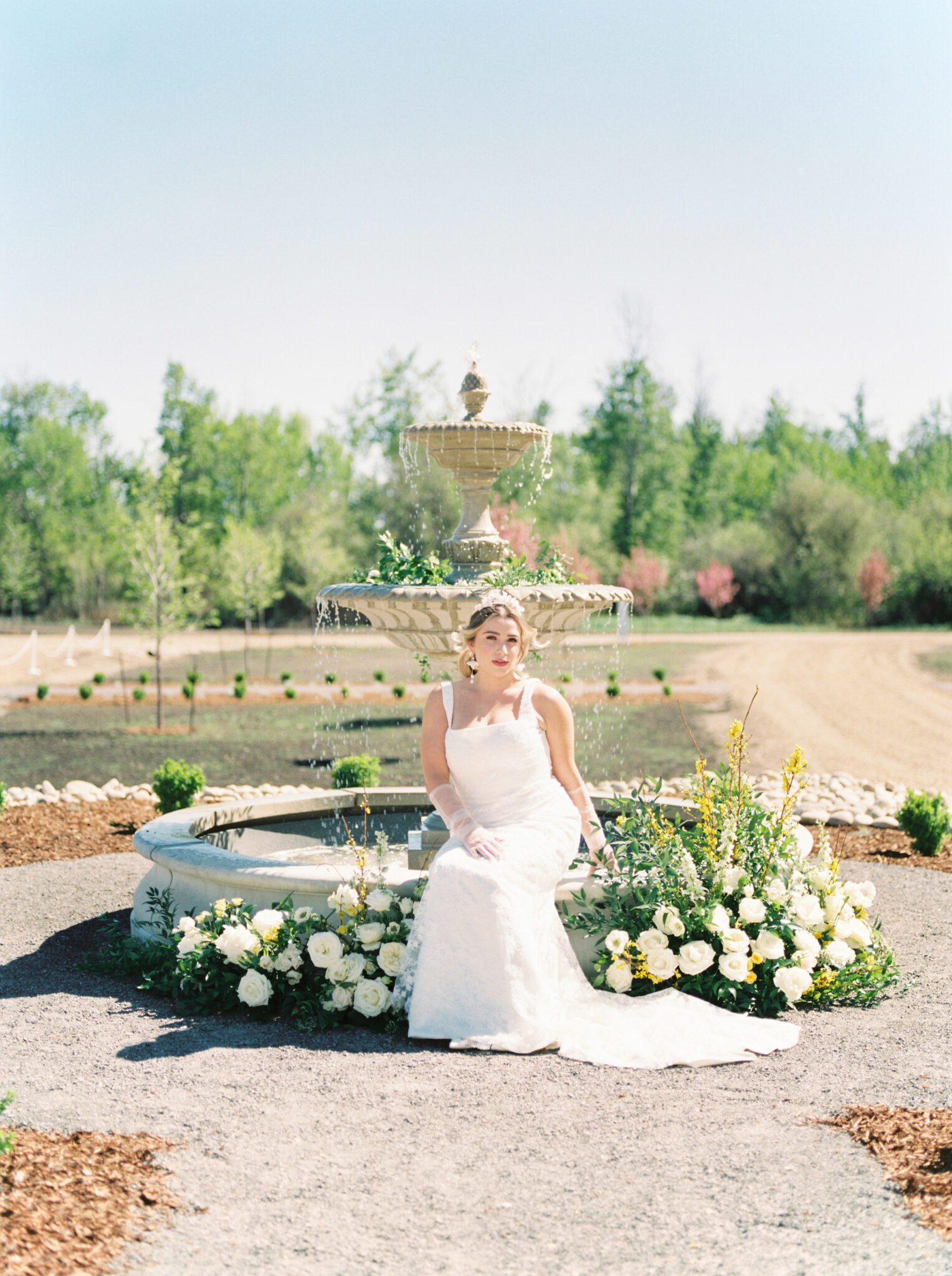 Bride sitting on the elegant water fountain at Sparrow Lane Events, surrounded by lush pale yellow and ivory florals by Lovella Lifestyle. Spring wedding inspiration, bridal fashion inspiration, sophisticated lace gown and sheer bridal gloves. 