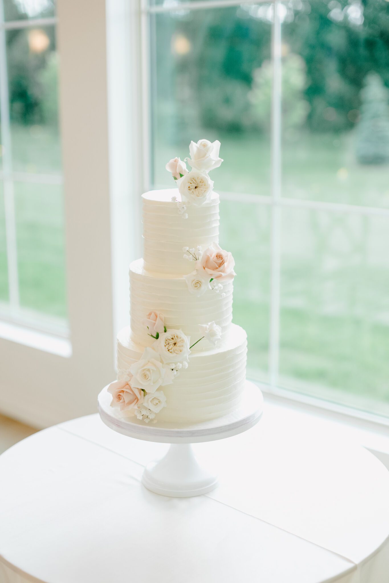 Beautiful classic three-tiered white wedding cake adorned with white and blush roses, created by Brianne Gabrielle Cakes.