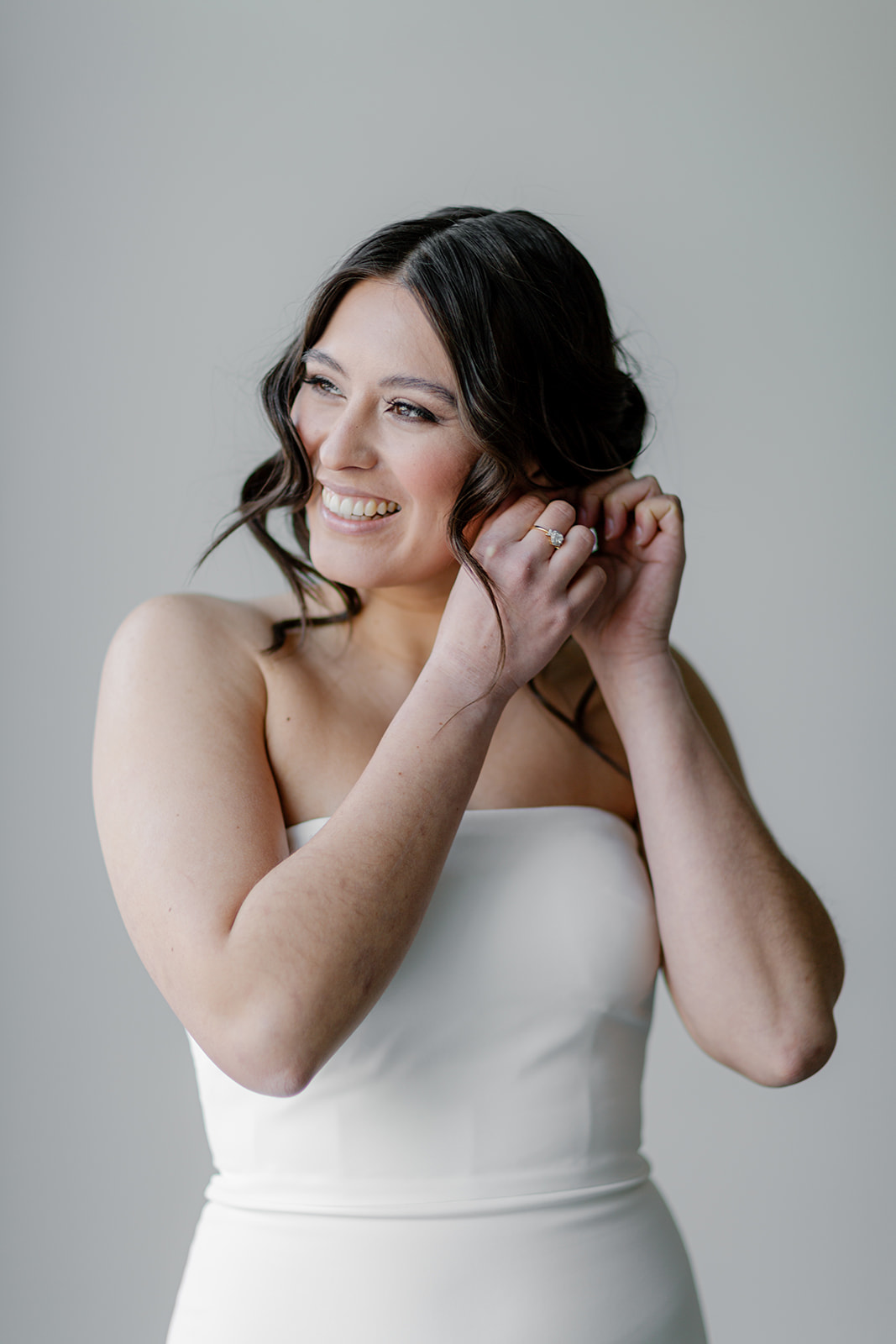 Gorgeous contemporary bride putting on earrings, wearing simple sleek wedding gown by Theia.