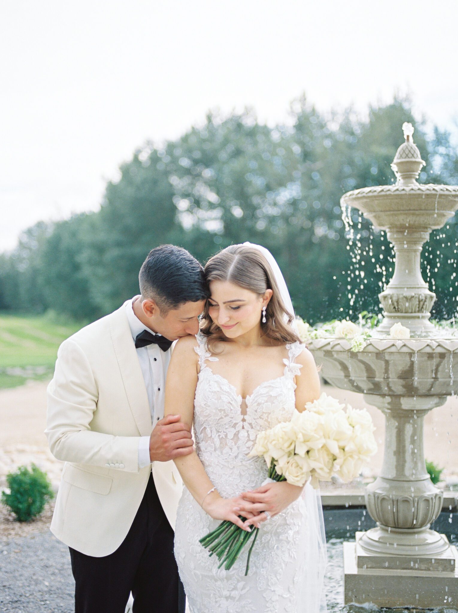 Elegant bride and groom embracing in front of water fountain at Sparrow Lane Events in Alberta. Classic colour palette of white, blush, and green wedding inspiration.