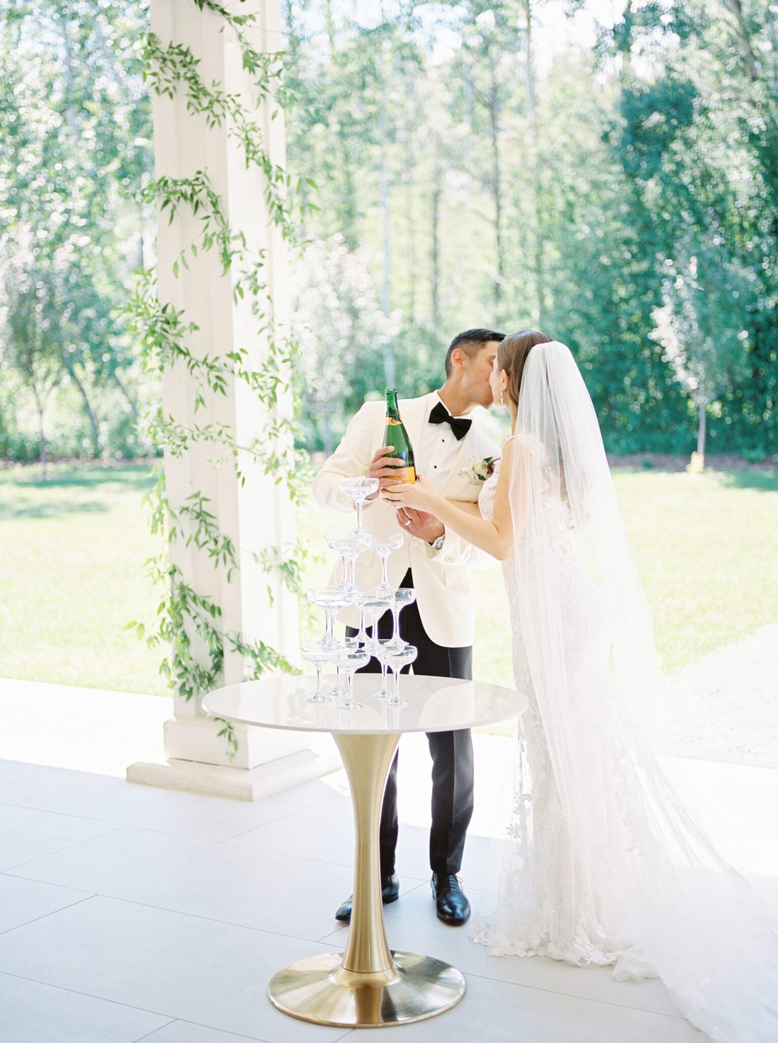 Couple popping champagne at Timeless Summer Wedding reception at Sparrow Lane Events in Alberta. Summer wedding features white roses, greenery blush decor accents and warm wood tones.