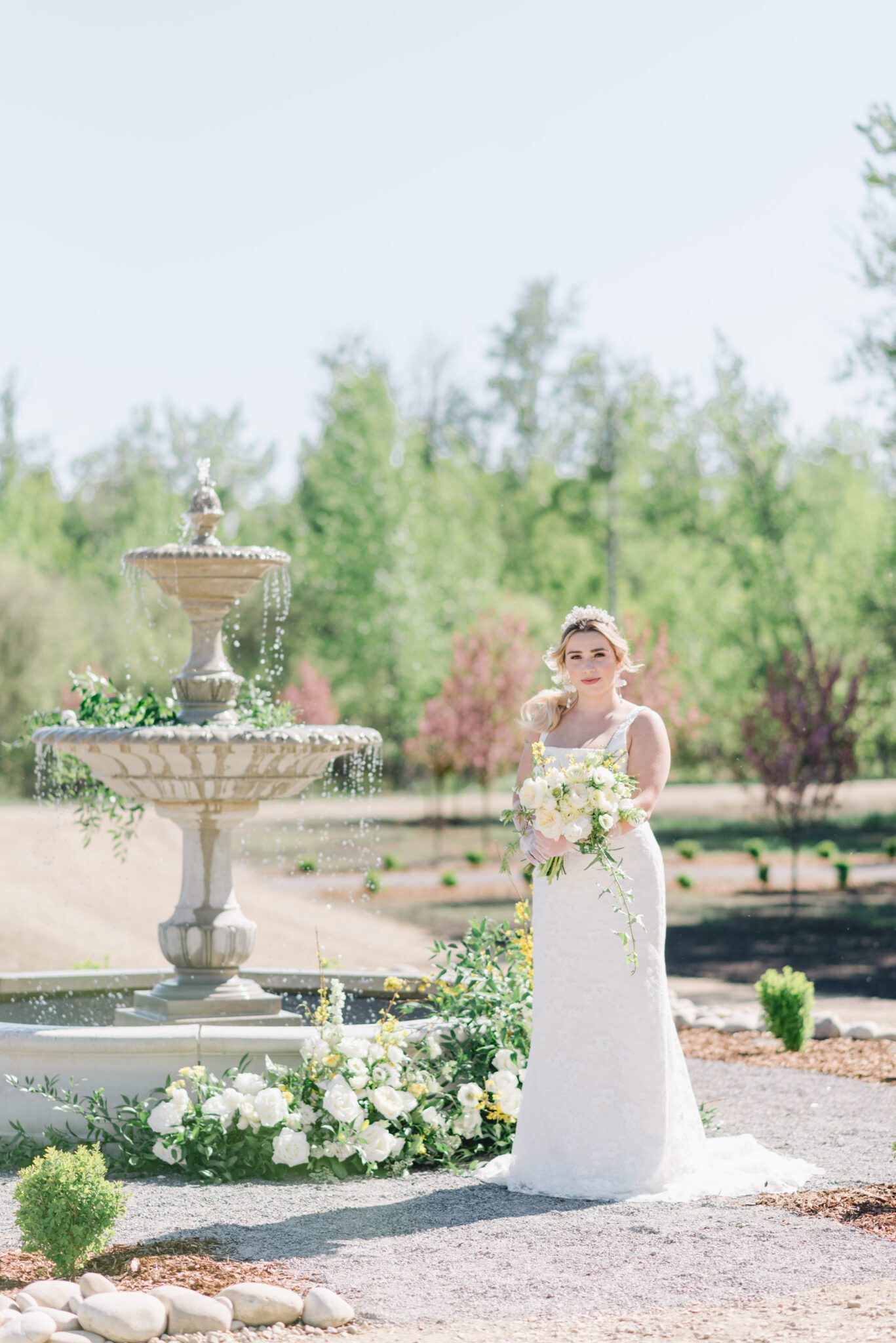 Bride standing in front of the water fountain at Sparrow Lane Events,  holding pale yellow, ivory and white floral bouquet of roses and peonies for the perfect spring wedding inspiration. 
