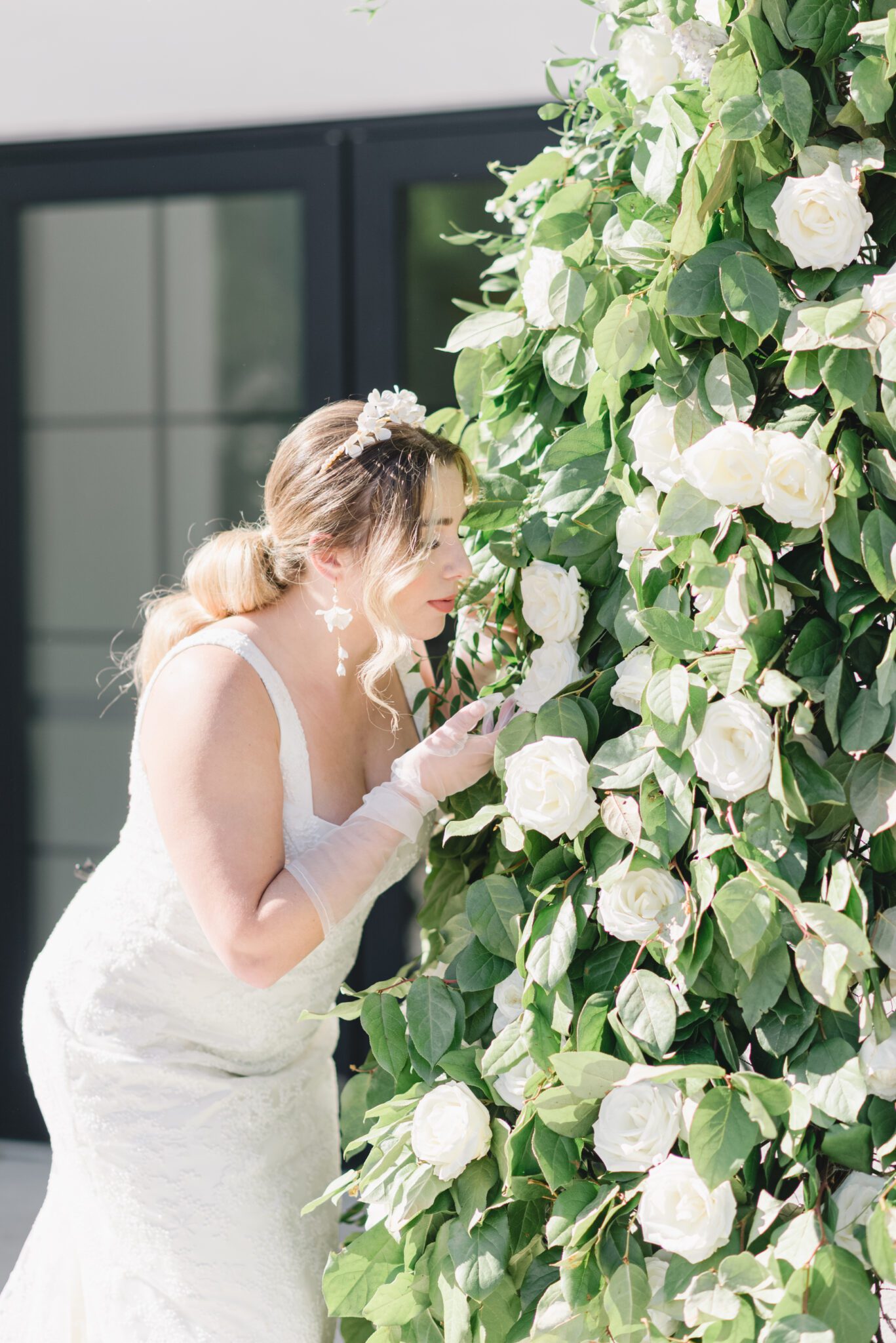 Bride standing in front of lush ivory, white and greenery floral installation wearing sophisticated yet whimsical bridal gown and jewelry. Outdoor summer wedding inspiration.