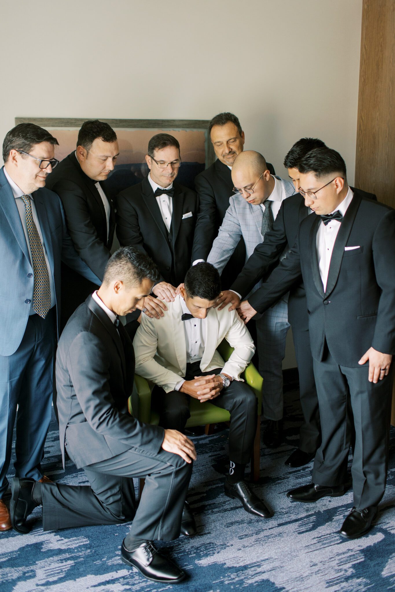 Groom surrounded by groomsmen and parents praying together on wedding day.