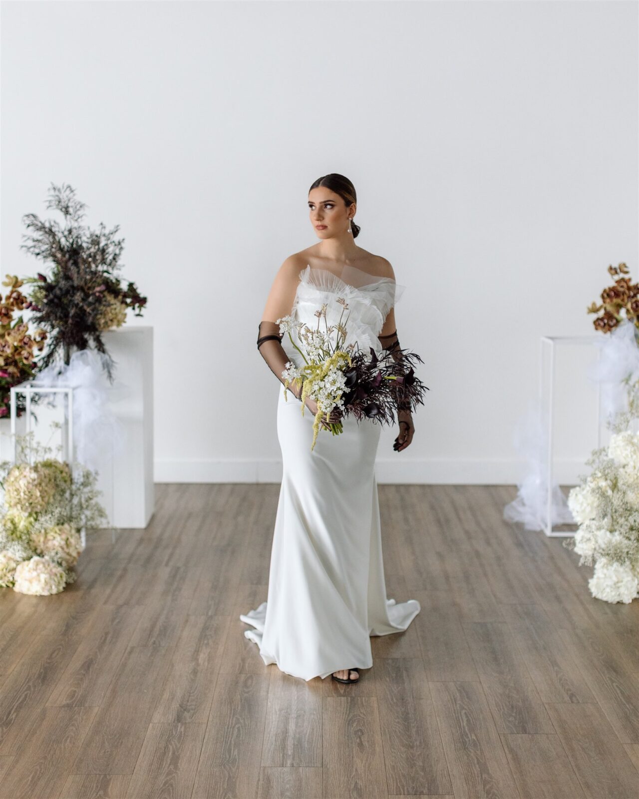 Modern bride wearing elegant, fit-and-flare strapless gown with an open back secured with a cross-strap. Architectural ruffles decorate the bodice while the crepe skirt flows out into a train by OUMA Bridal.