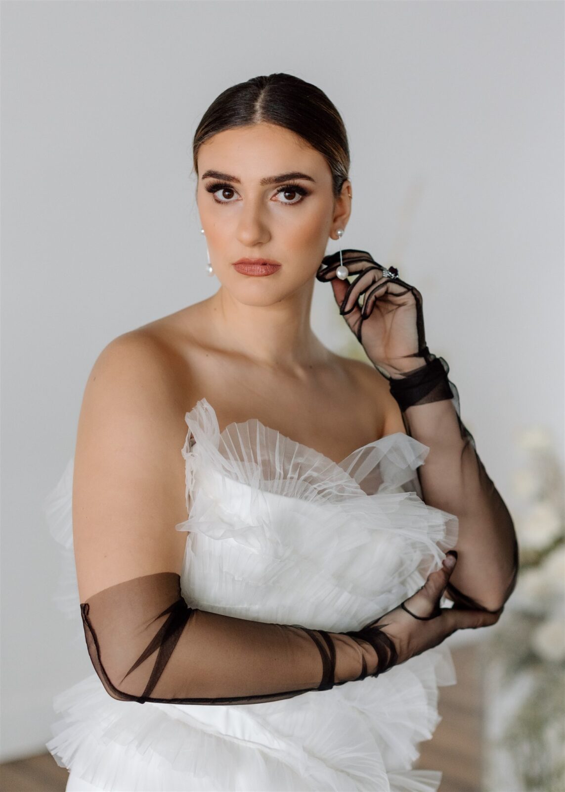 Modern bride wearing elegant, fit-and-flare strapless gown and sheer black bridal gloves. Architectural ruffles decorate the bodice while the crepe skirt flows out into a train by OUMA Bridal.