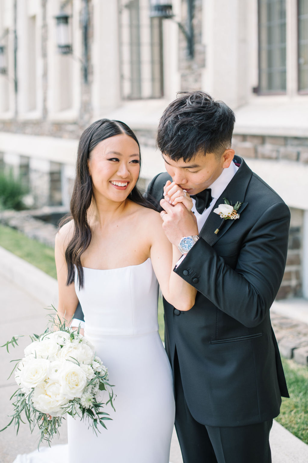 Bride and groom at their Fairmont Banff Springs Golf and Country Club wedding. Bride wearing sleek form fitting white bridal gown, holding classic bouquet of white roses and greenery. Summer wedding inspiration in Banff, Alberta. 