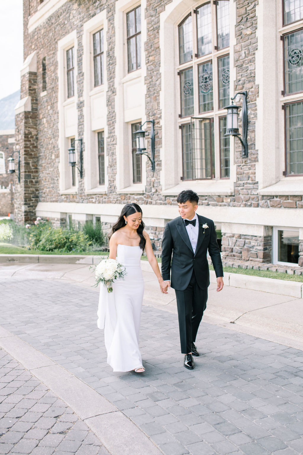 Bride and groom at their Fairmont Banff Springs Golf and Country Club wedding. Bride wearing sleek form fitting white bridal gown, holding classic bouquet of white roses and greenery. Summer wedding inspiration in Banff, Alberta. 