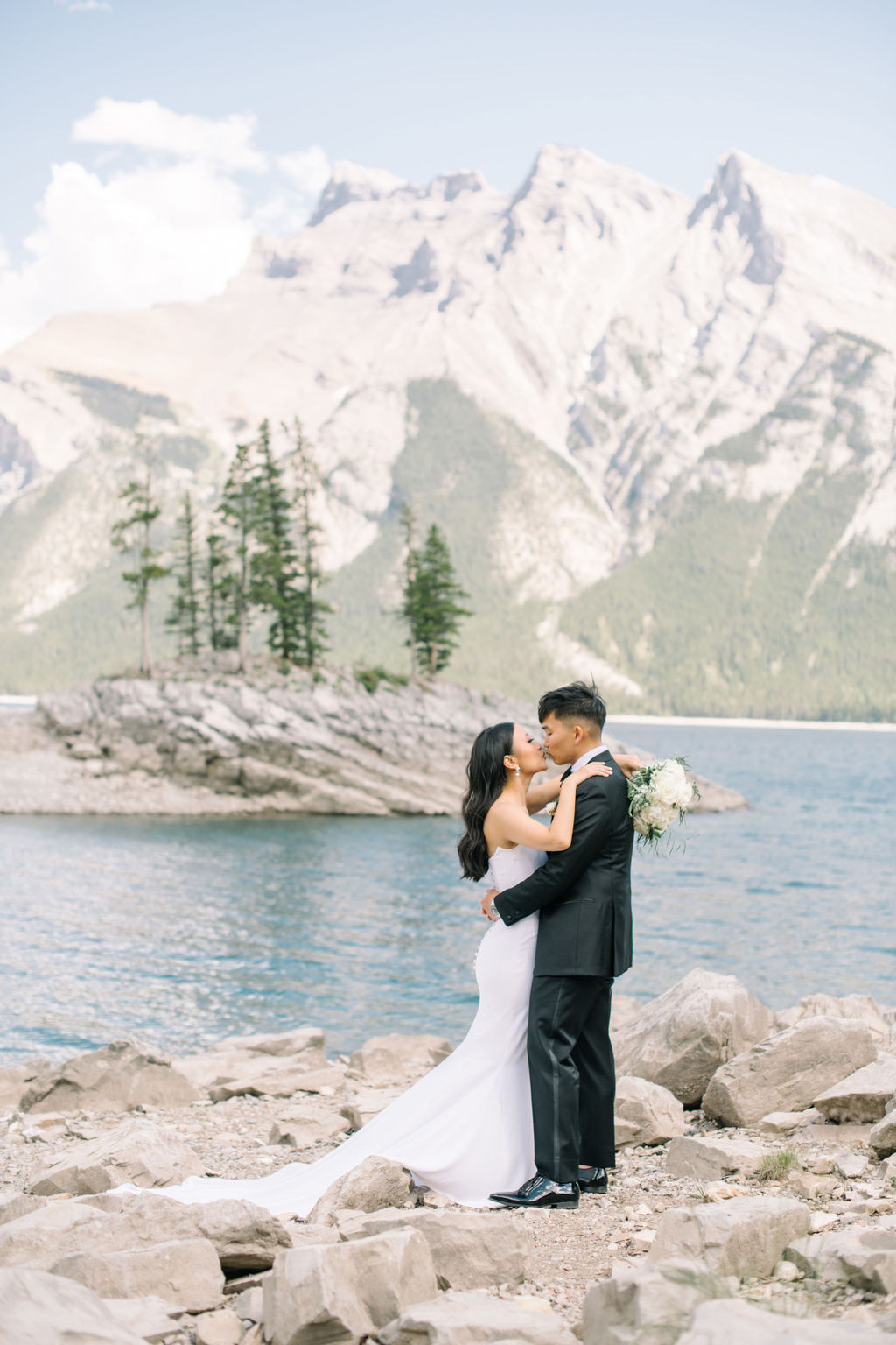 Elegant bride and groom embracing in the mountains of Banff, Alberta with lake in the background. Canadian summer mountain wedding inspiration. Sophisticated mountain bridal portraits by Corrina Walker Photography. 