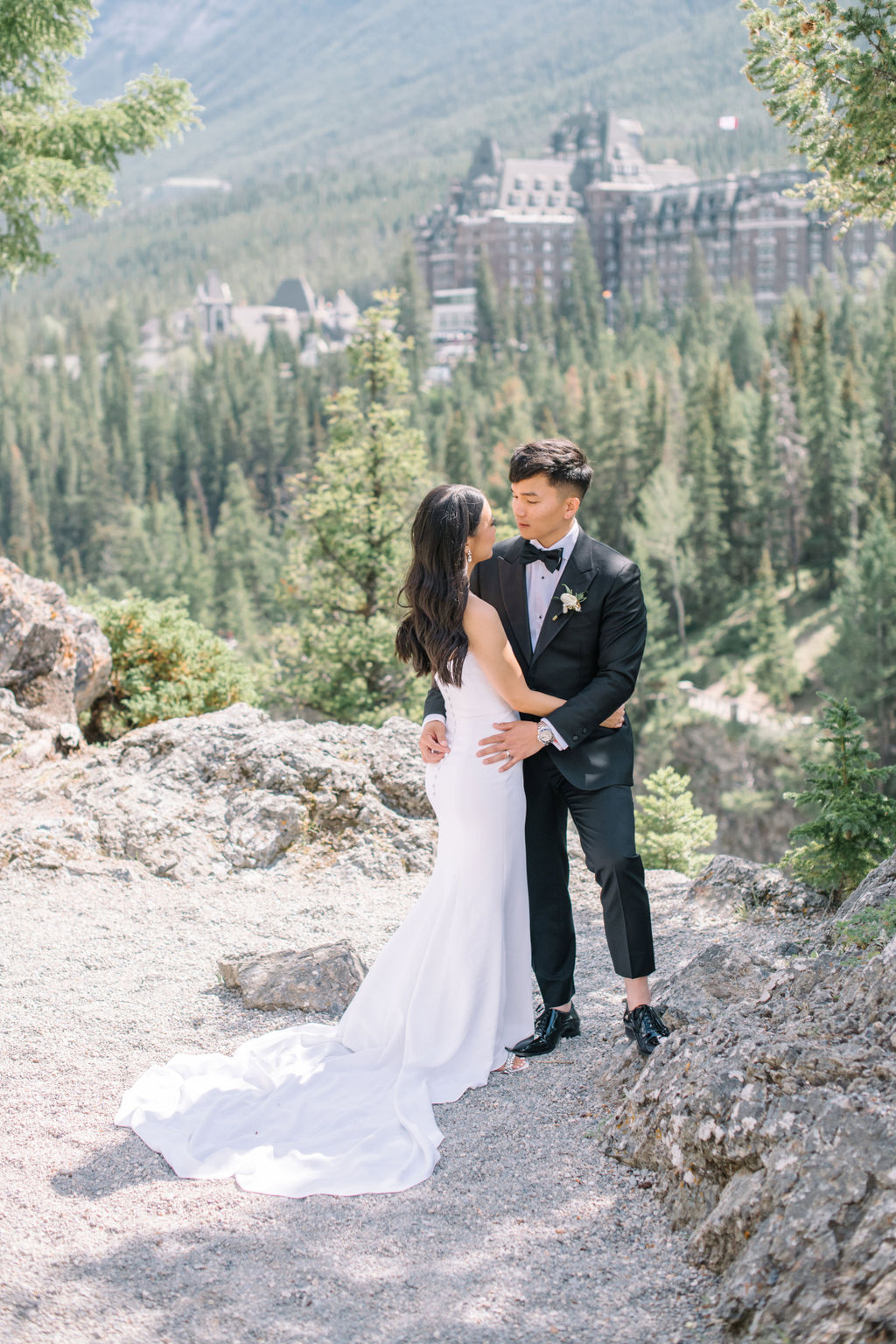 Elegant bride and groom embracing in the mountains with Fairmont Banff Springs Golf and Country Club in the background.  Canadian summer mountain wedding inspiration.