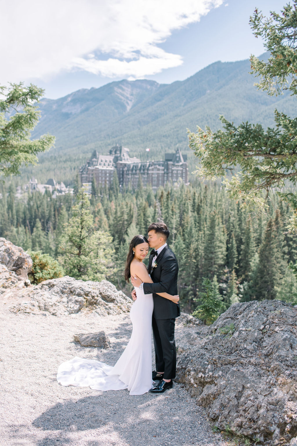 Elegant bride and groom embracing in the mountains with Fairmont Banff Springs Golf and Country Club in the background.  Canadian summer mountain wedding inspiration.