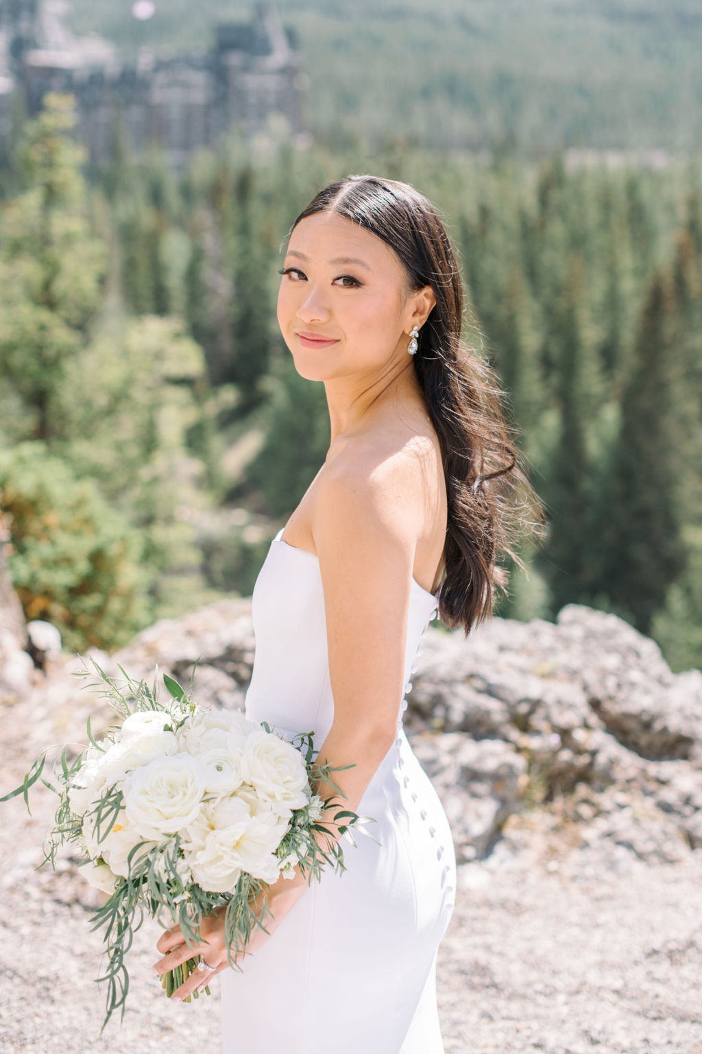 Bride wearing sleek form fitting white bridal gown, holding classic bouquet of white roses and greenery. Summer wedding inspiration in Banff, Alberta. 