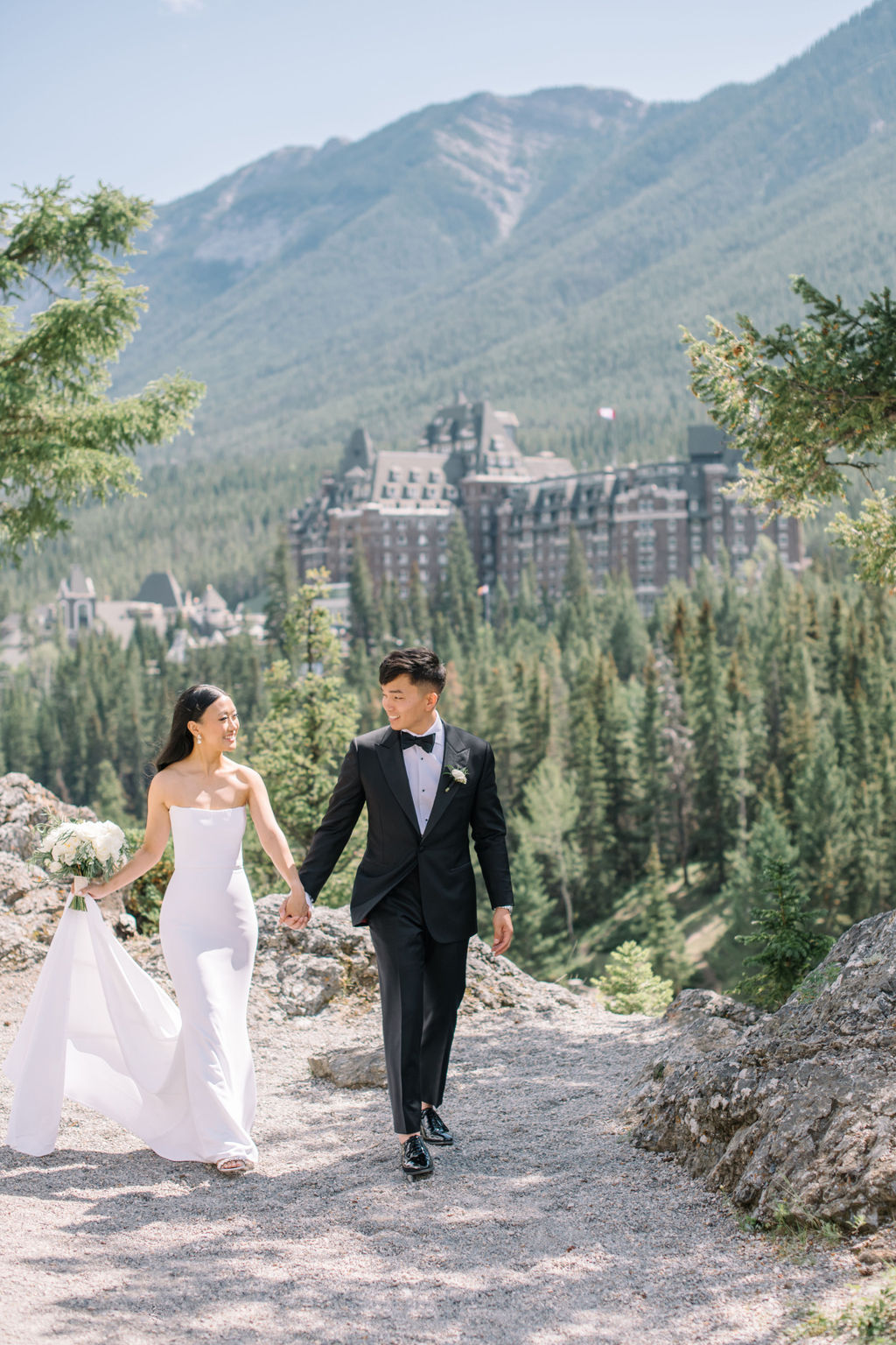 Elegant bride and groom walking holding hands in the mountains with Fairmont Banff Springs Golf and Country Club in the background.  Canadian summer mountain wedding inspiration.