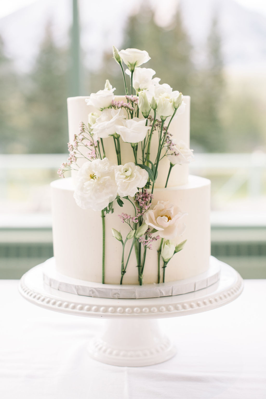 Classic two-tiered white wedding cake by Pretty Sweet YYC decorated with delicate white and pink florals. Simple and elegant summer wedding cake inspiration. 