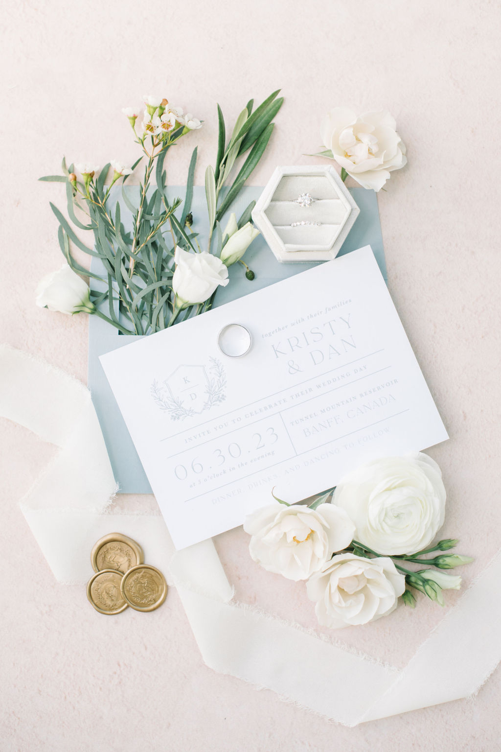 Elegant white and dusty blue wedding invitation paper suite styled with white roses and greenery, gold wax envelope seals, blush background, white raw silk ribbon, photographed by Corrina Walker Photography. 
