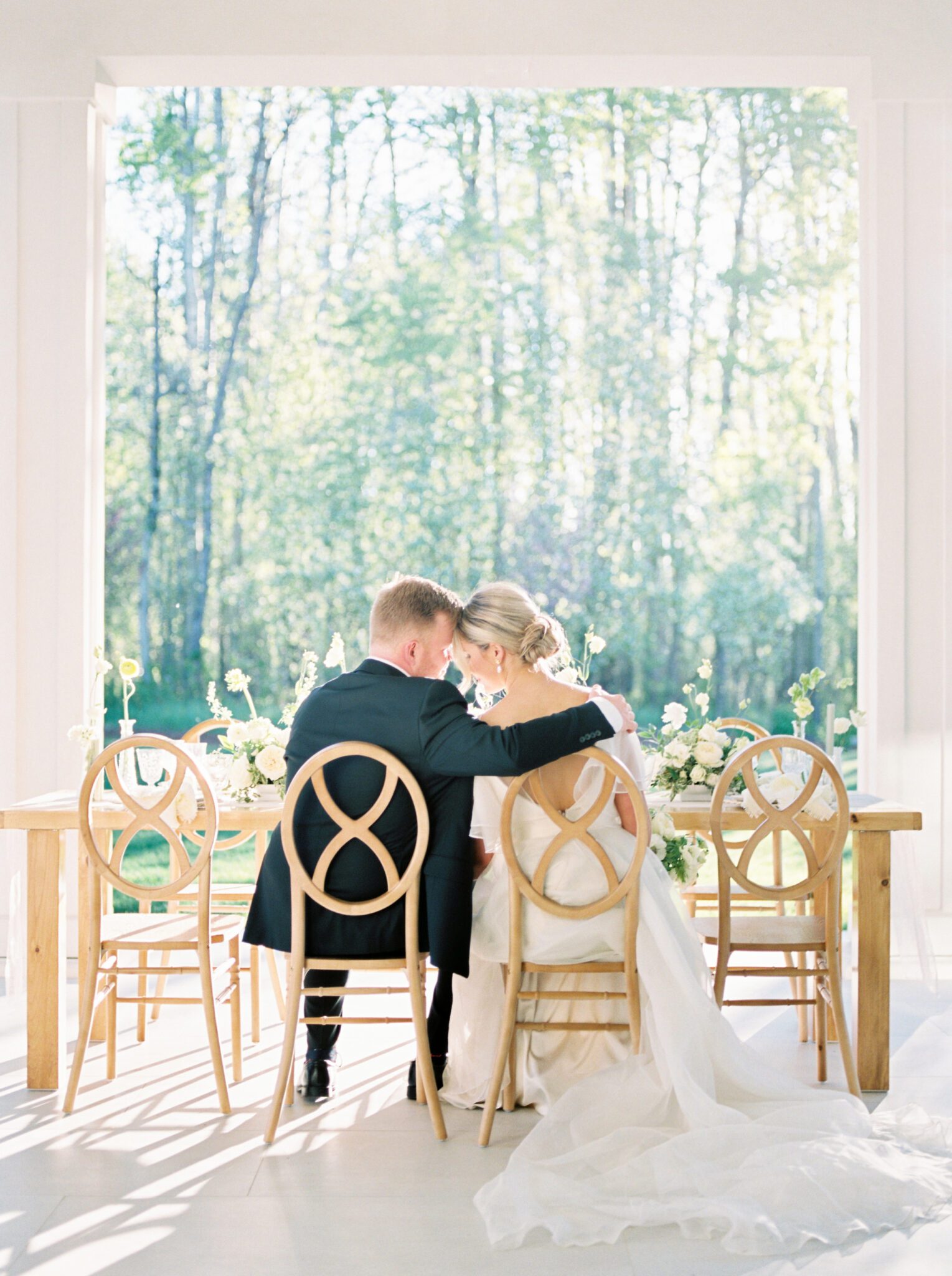 Couple sitting at cottage core inspired wedding reception at Sparrow Lane Weddings and Events venue near Edmonton, Alberta. Fine art summer wedding. Wooden infinity chairs, outdoor summer wedding tablescape with lush florals in pale yellow, ivory and white with greenery. 