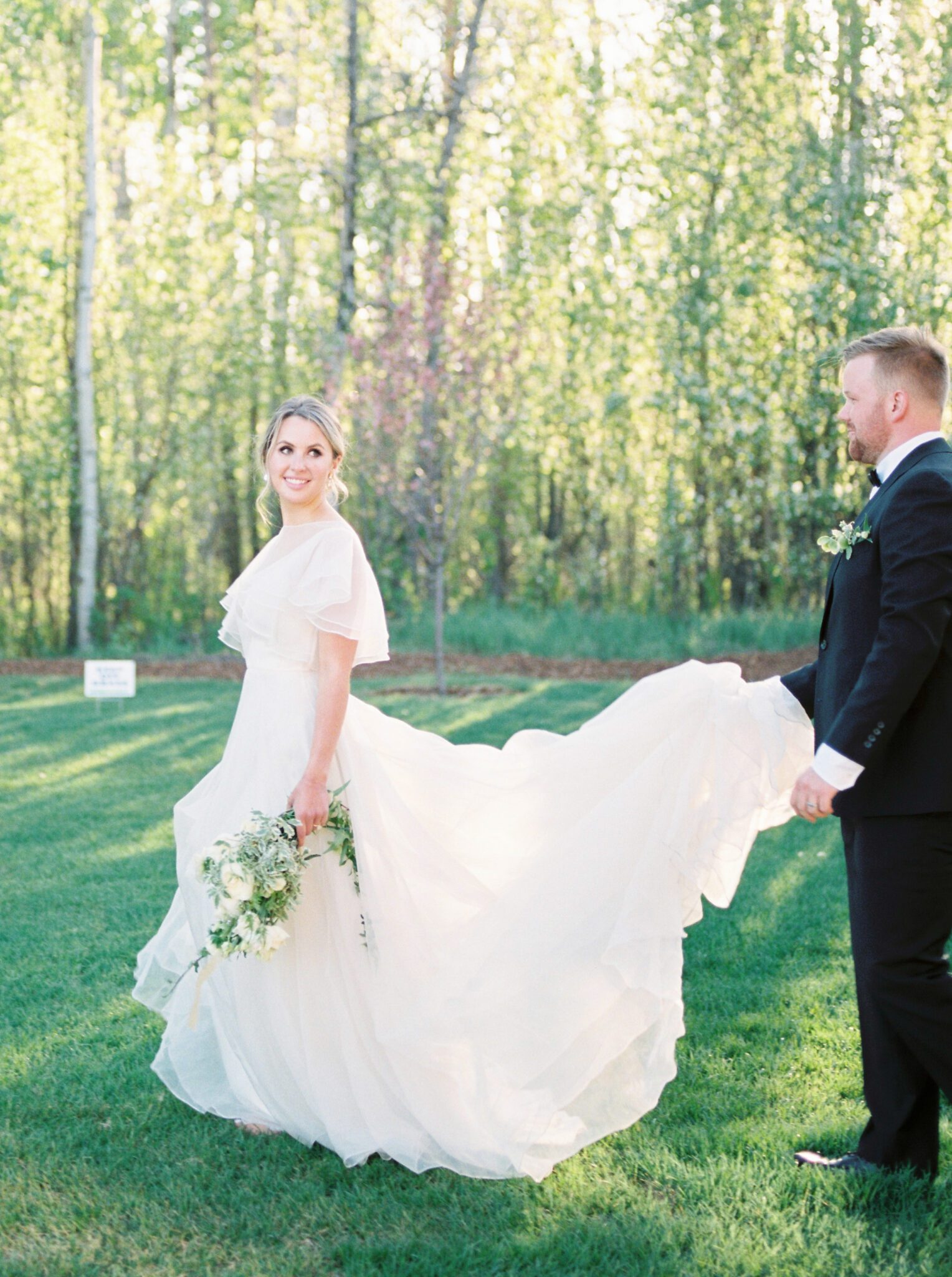 Bride and groom embracing in front of Sparrow Lane in Edmonton, Alberta during fine art summer wedding. Pale yellow, ivory, white and green cottage core inspired wedding inspiration. Bride wearing whimsical yet sophisticated bridal gown.