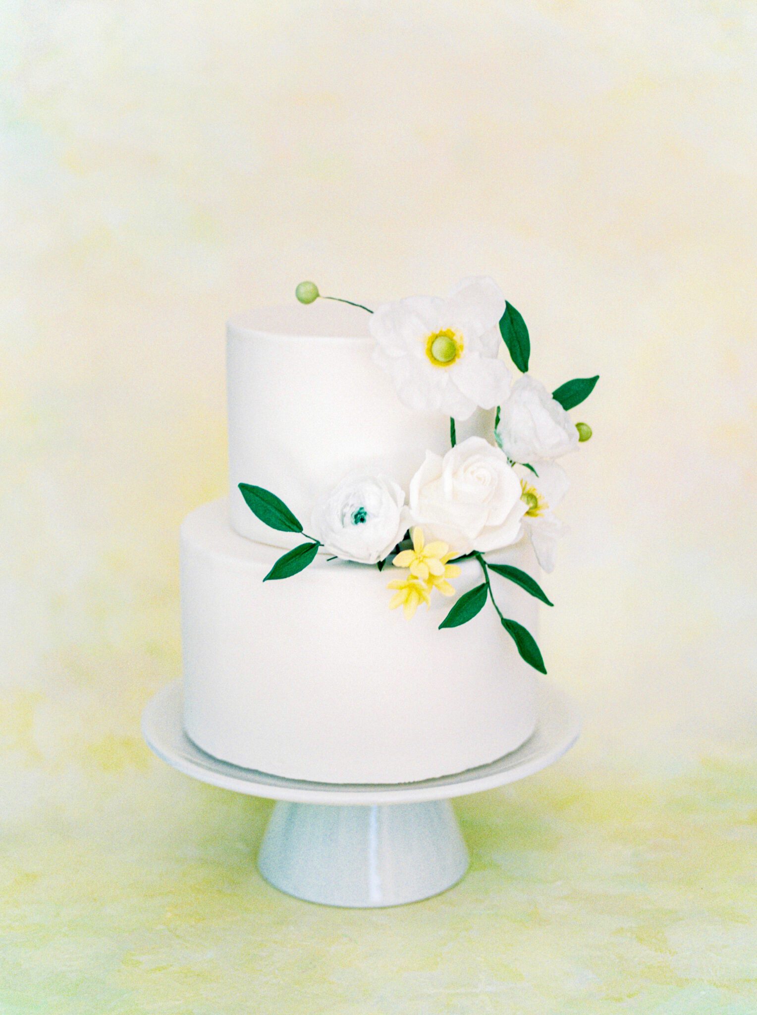 Elegant cottage core inspired 3-tiered white wedding cake decorated with yellow and white florals and greenery by Brianne Gabrielle Cakes. Spring wedding cake inspiration.