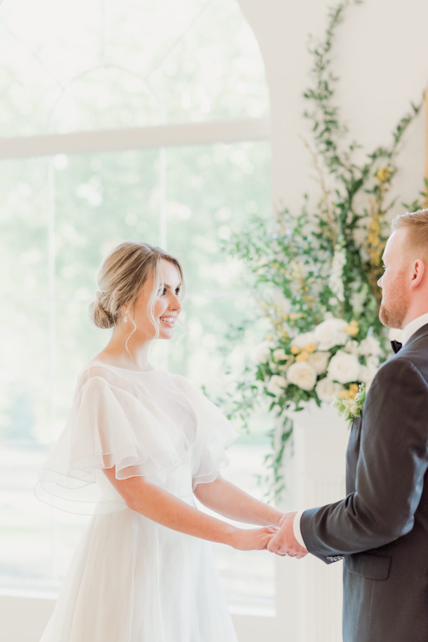 Bride and groom during fine art Indoor wedding ceremony at Sparrow Lane with a lush floral installation climbing up the fireplace. Pale yellow, ivory, white, colour palette. Bride wearing a whimsical yet sophisticated bridal gown.