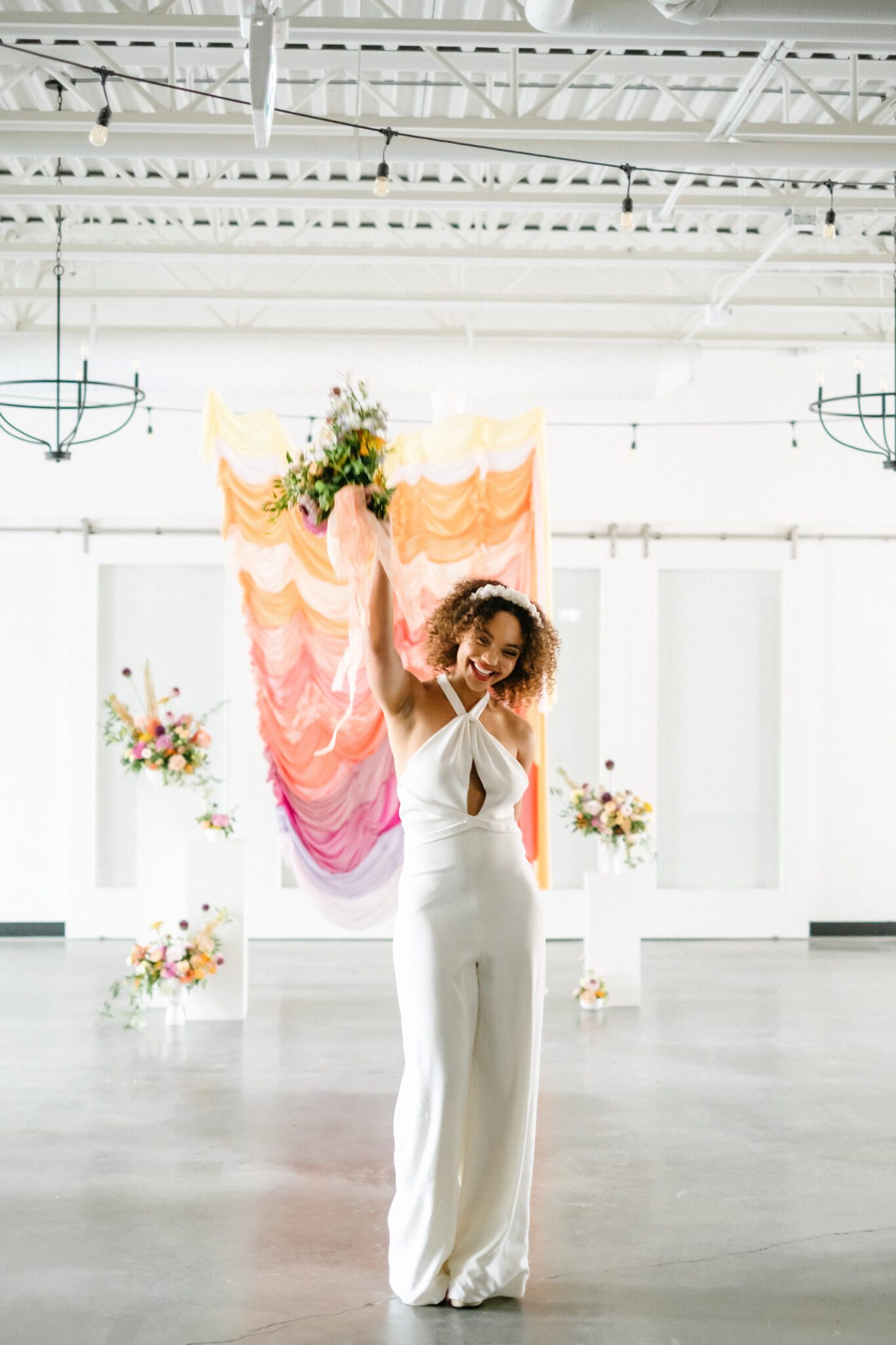Yellow, tangerine, peach, pink, and purple ruched fabric ceremony backdrop, featuring whimsical florals by Meadow & Vine at The Brownstone in Calgary, Alberta. Bride wearing retro-inspired bridal jumpsuit and custom floral headpiece.