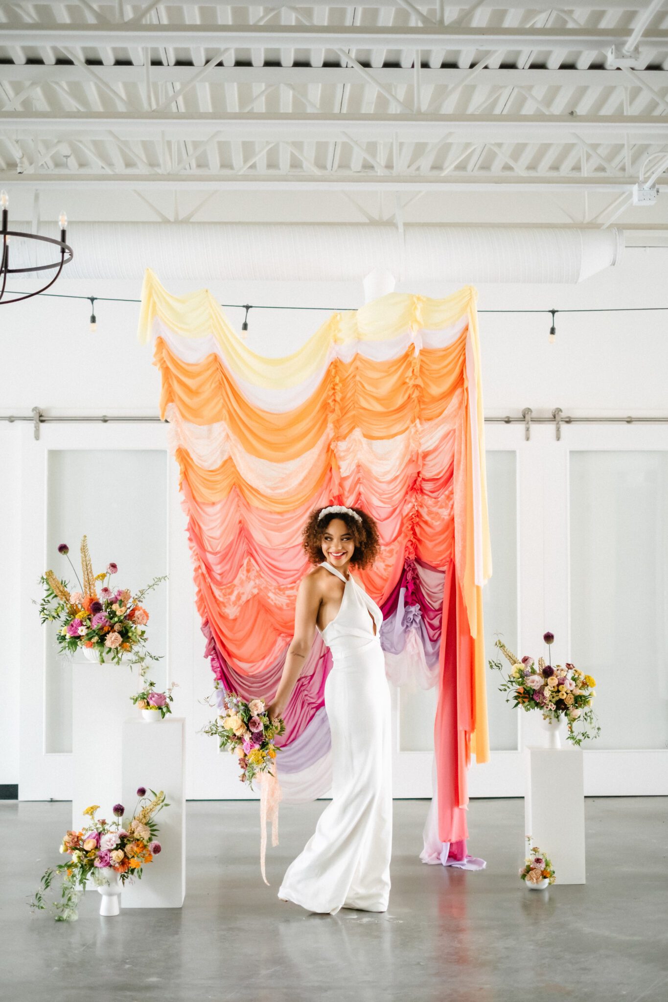 Yellow, tangerine, peach, pink, and purple ruched fabric ceremony backdrop, featuring whimsical florals by Meadow & Vine at The Brownstone in Calgary, Alberta. Bride wearing retro-inspired bridal jumpsuit and custom floral headpiece.
