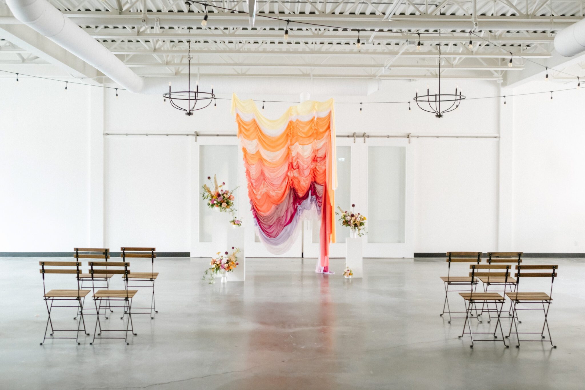 Yellow, tangerine, peach, pink, and purple ruched fabric ceremony backdrop, featuring whimsical florals by Meadow & Vine at The Brownstone in Calgary, Alberta. 