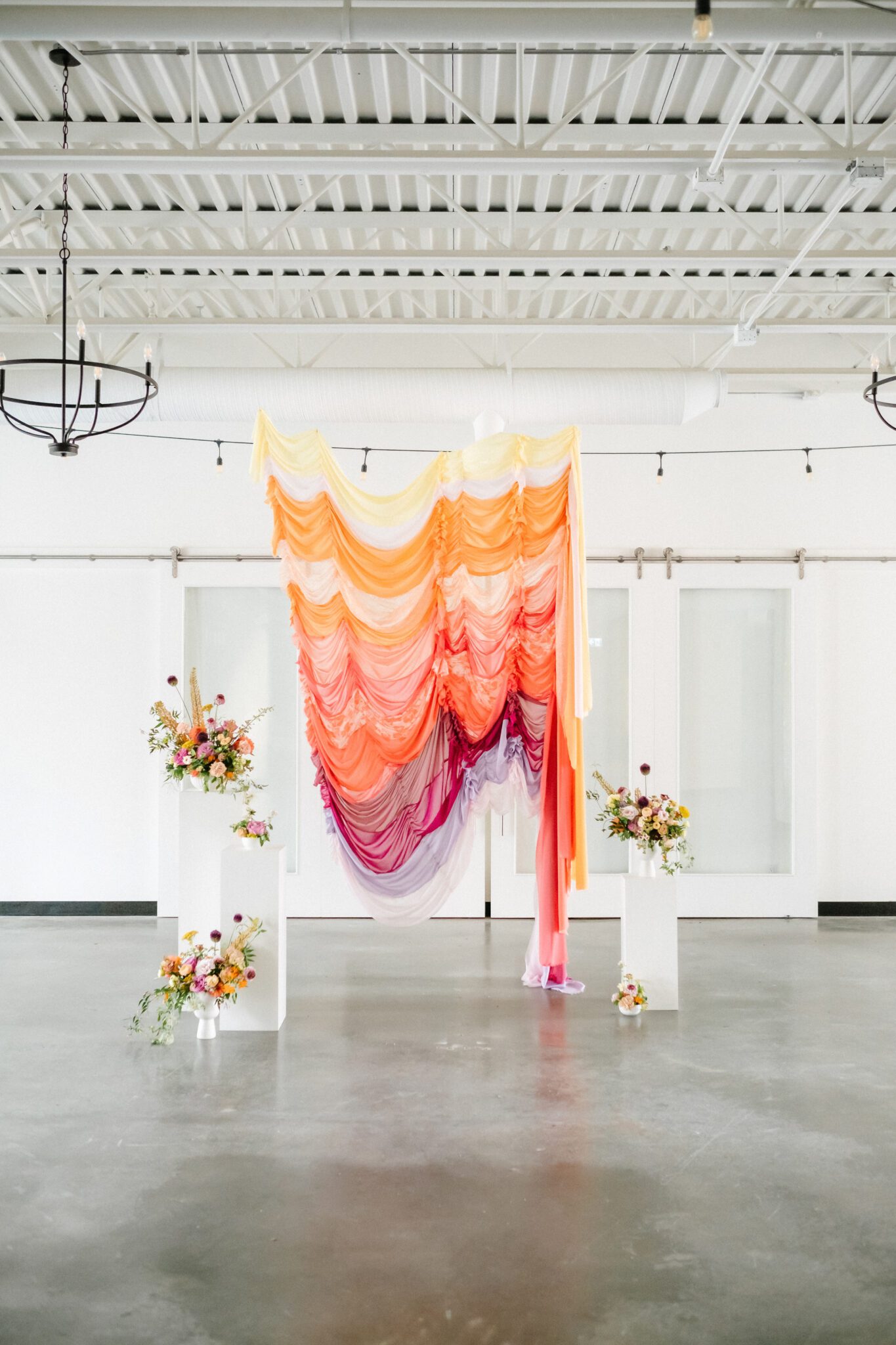 Yellow, tangerine, peach, pink, and purple ruched fabric ceremony backdrop, featuring whimsical florals by Meadow & Vine at The Brownstone in Calgary, Alberta. 