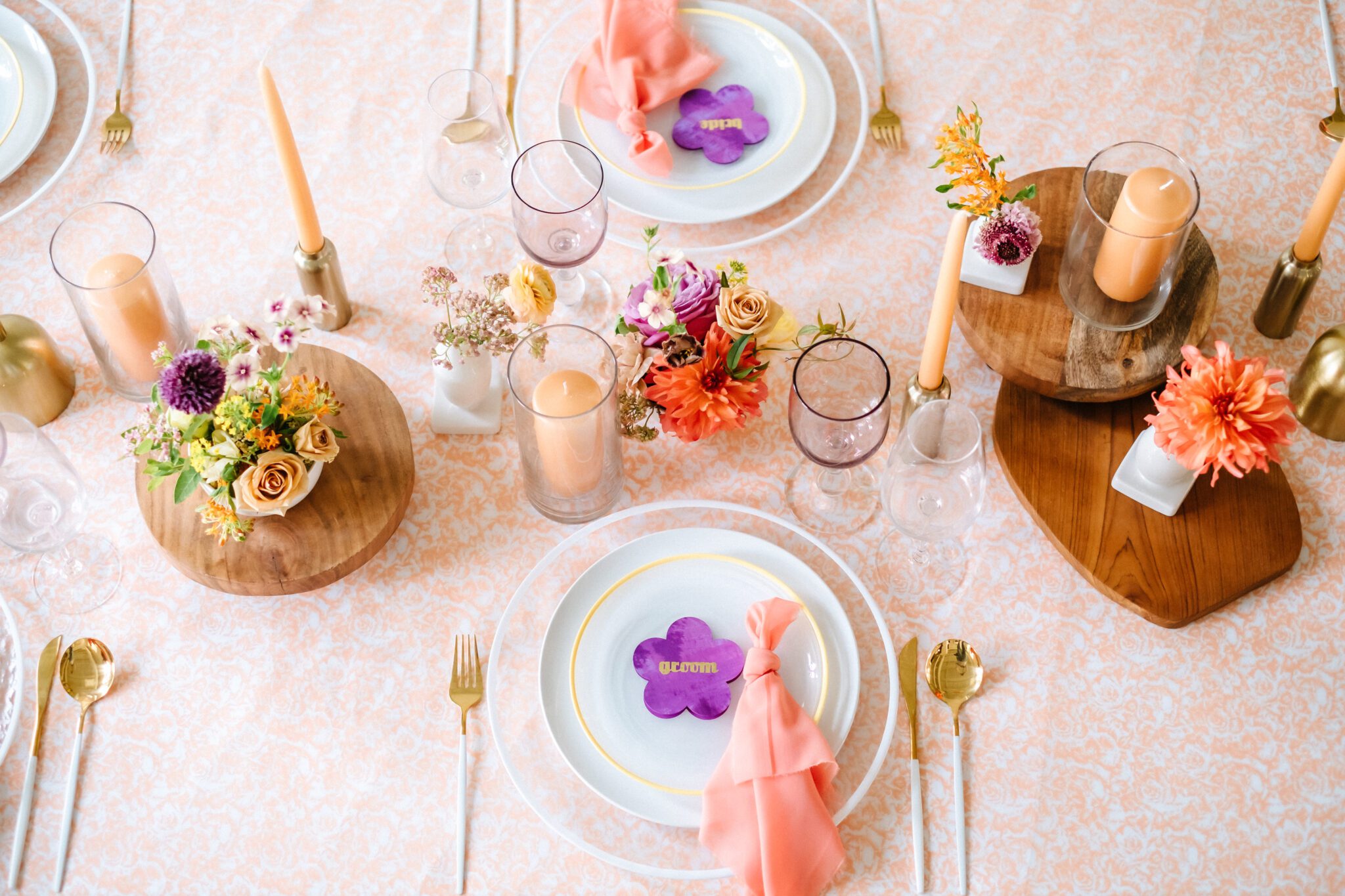 Bright & colourful brunch wedding tablescape. Florals by Meadow & Vine. Retro-inspired vibrant colour palette of purple, orange, yellow, peach, tangerine and coral. Wood and gold accents. Custom retro purple flower name place card. 