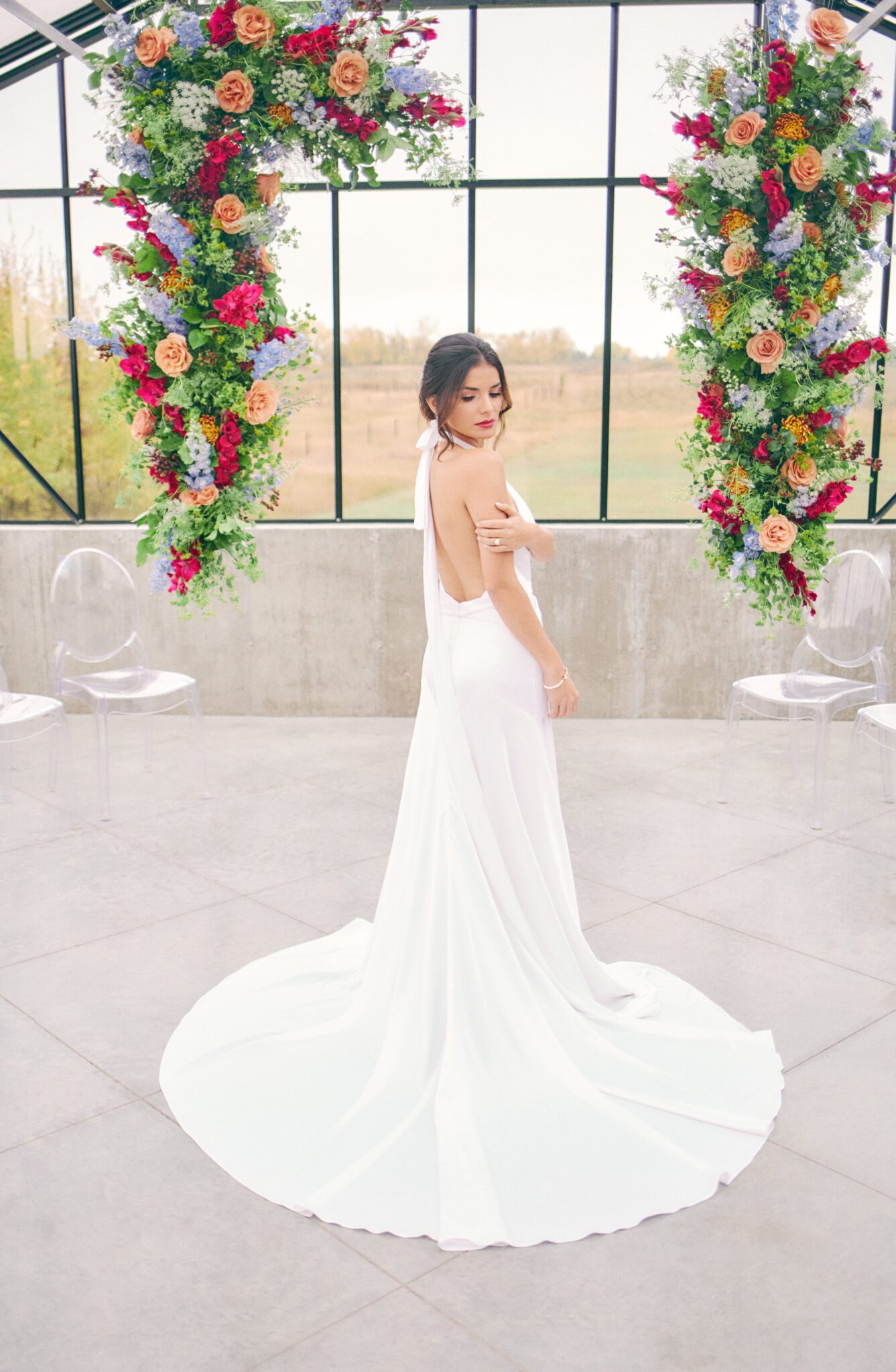 Bride standing in front of dramatic and colourful pink, peach, orange, and violet floating floral arch framing intimate ceremony space at The Station in Saskatchewan. Vibrant and bold wedding inspiration.