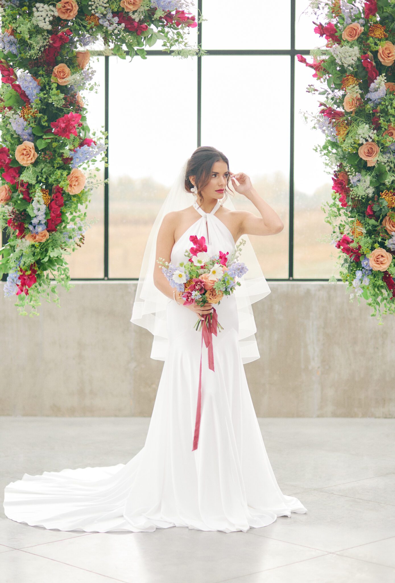 Bride standing in front of dramatic and colourful pink, peach, orange, and violet floating floral arch framing intimate ceremony space at The Station in Saskatchewan. Vibrant and bold wedding inspiration.