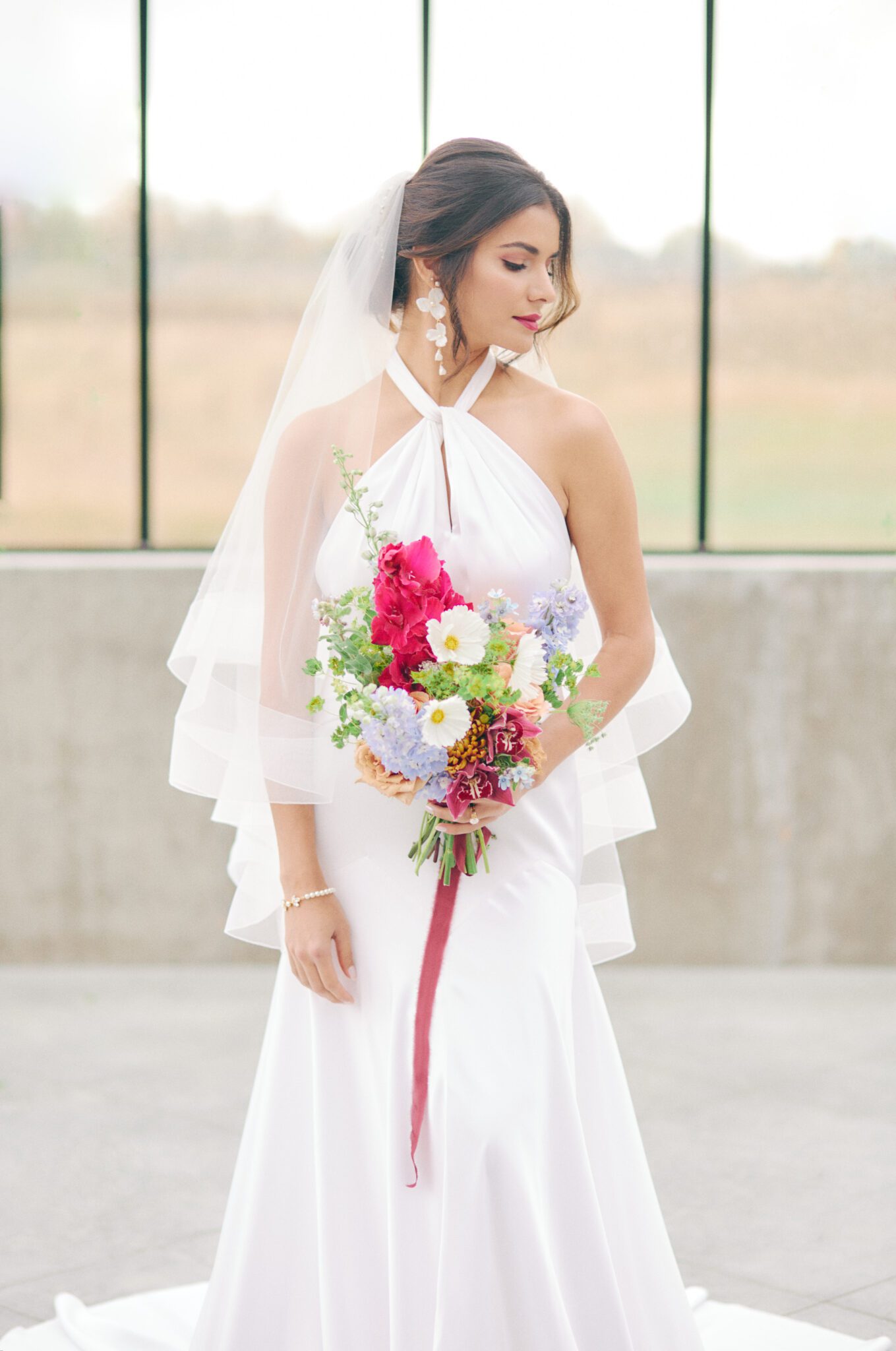 Elegant and modern bride wears a halter style wedding dress, pearl hairpiece, and statement earrings by Joanna Bisley Designs, and veil. Bridal fashion inspiration. 