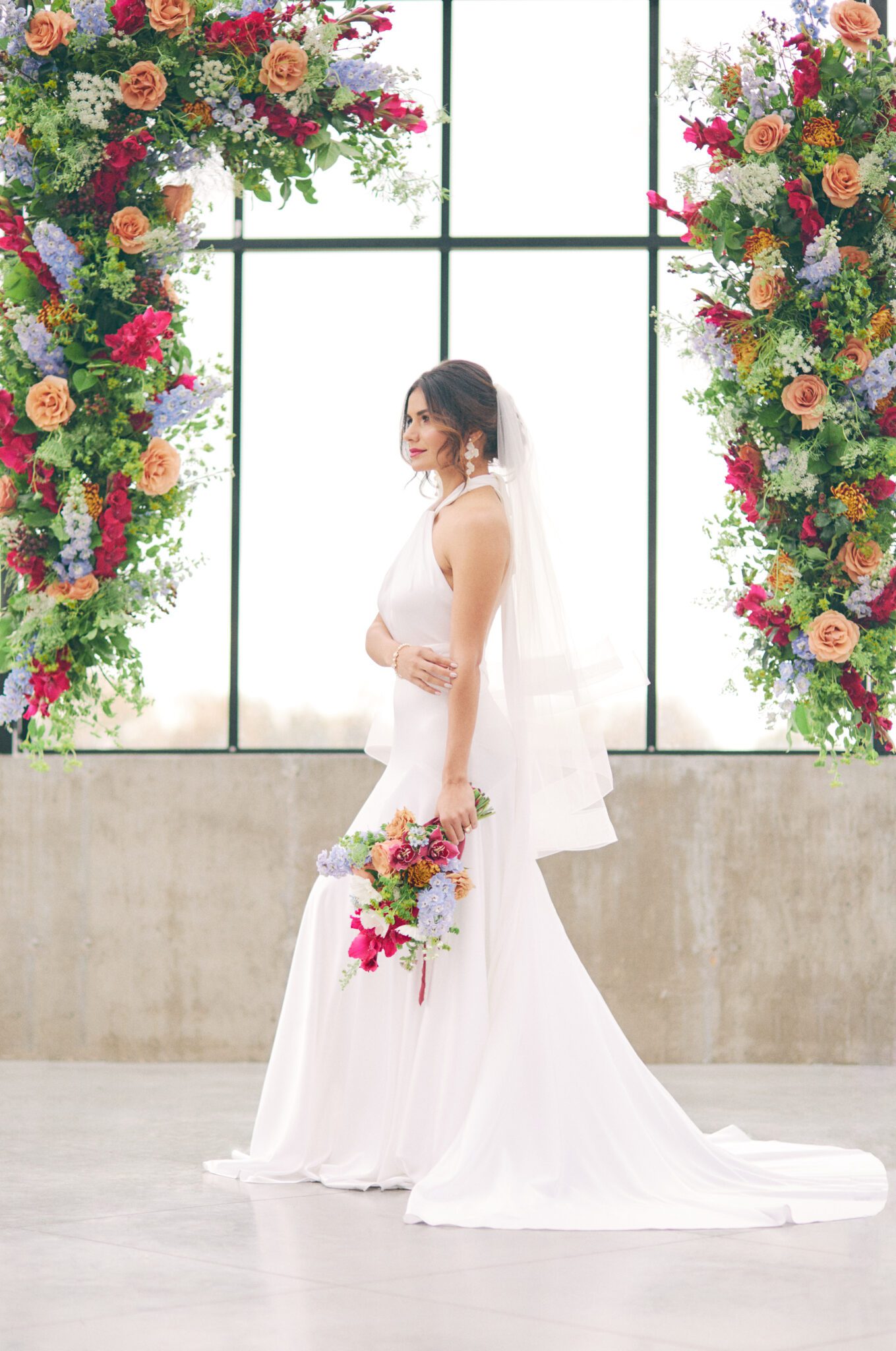 Bride standing in front of dramatic and colourful pink, peach, orange, and violet floating floral arch framing intimate ceremony space at The Station in Saskatchewan. Vibrant and bold wedding inspiration.