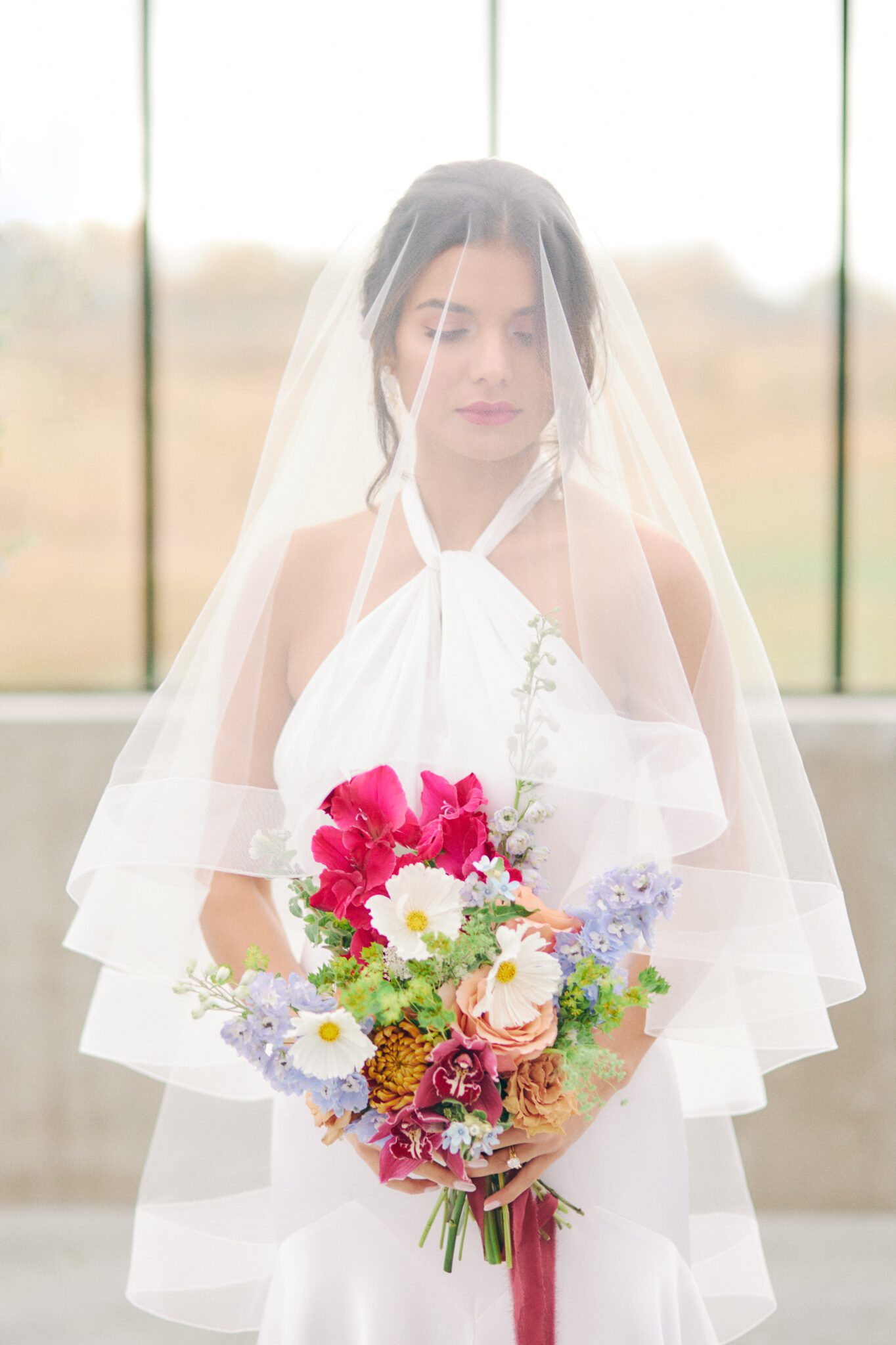 Elegant and modern bride wears a halter style wedding dress, pearl hairpiece, and statement earrings by Joanna Bisley Designs, and veil. Bridal fashion inspiration. Bold and vibrant wedding bouquet of hot pink, violet, orange, peach and burgundy. 