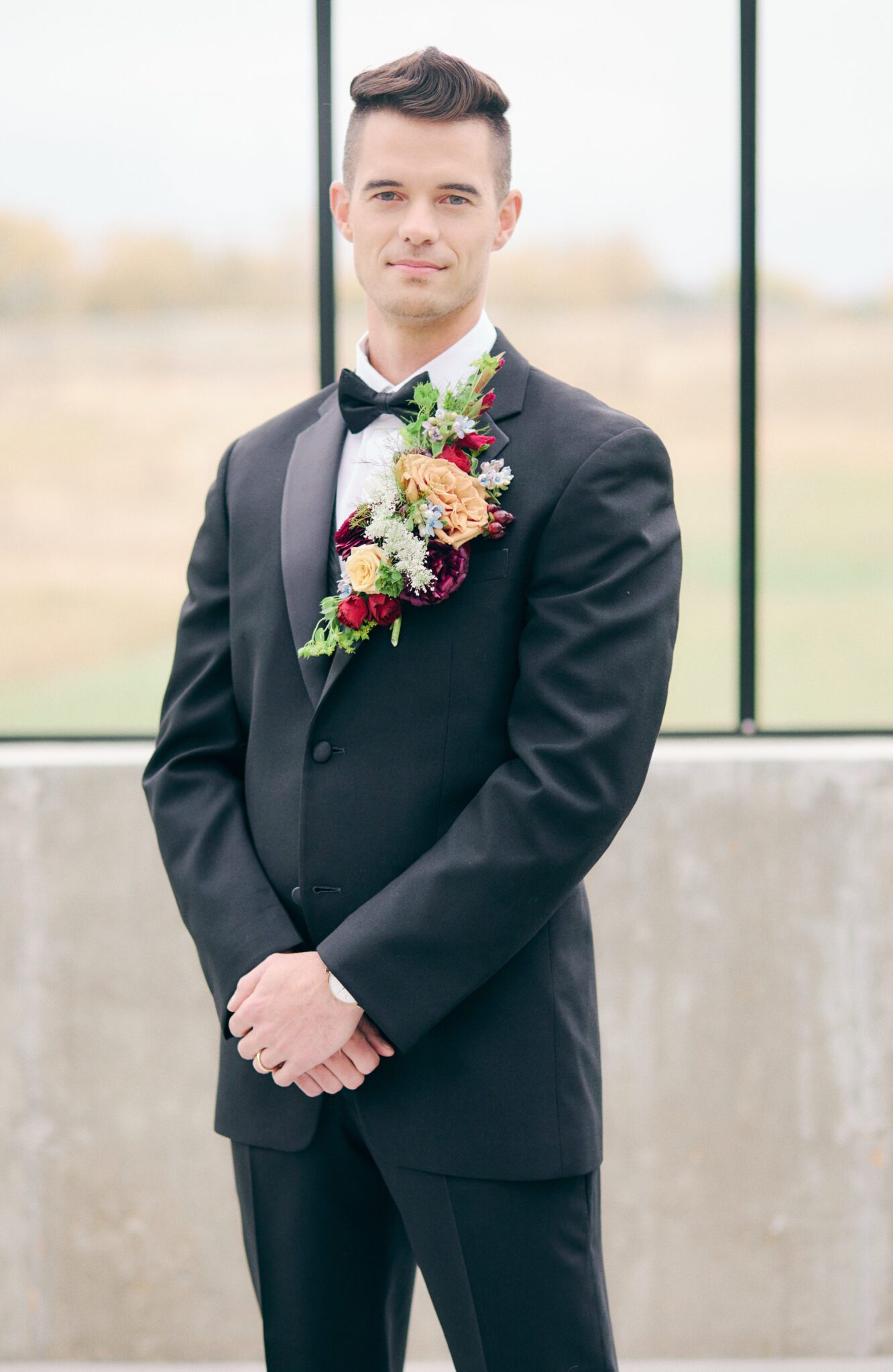 Elegant and modern groom wears classic black tuxedo with bold and vibrant cascading boutonniere of peach, pink, red, violet, and greenery. 
