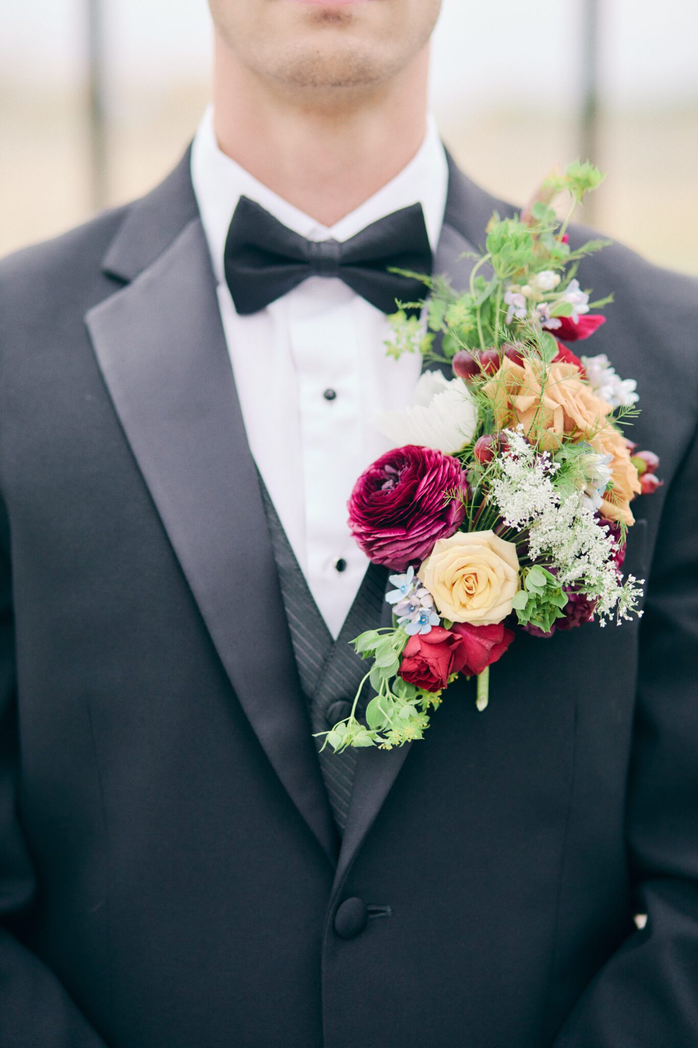 Elegant and modern groom wears classic black tuxedo with bold and vibrant cascading boutonniere of peach, pink, red, violet, and greenery. 
