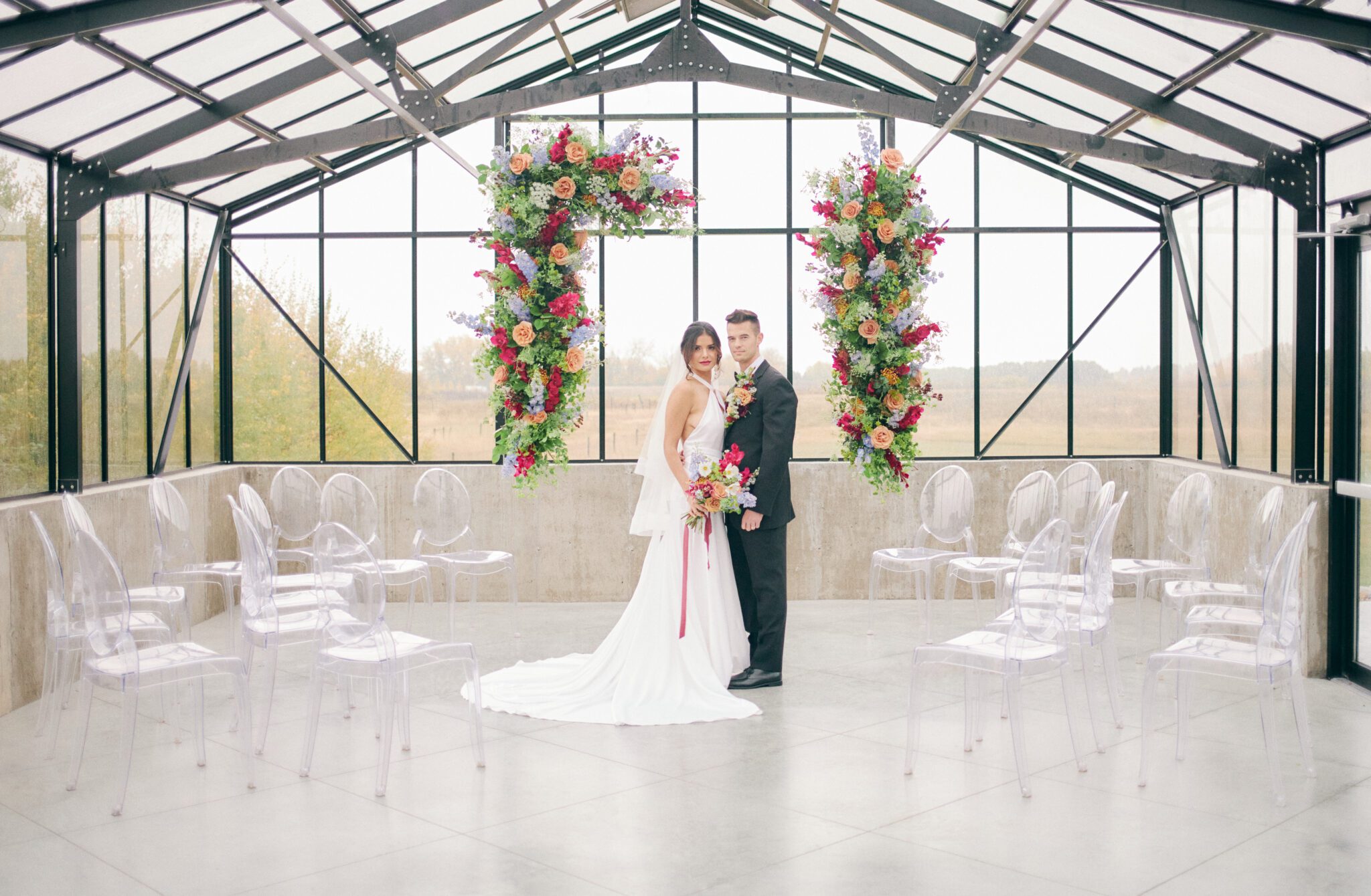 Bride and groom standing in front of dramatic and colourful pink, peach, orange, and violet floating floral arch framing intimate solarium ceremony space at The Station in Saskatchewan. Vibrant and bold wedding inspiration.