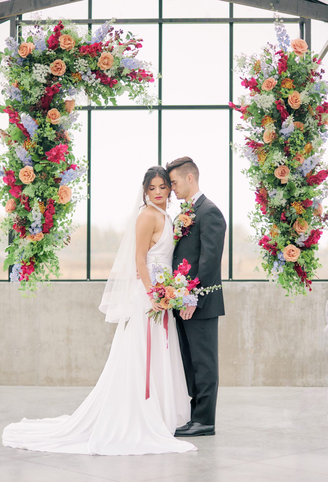 Bride and groom standing in front of dramatic and colourful pink, peach, orange, and violet floating floral arch framing intimate ceremony space at The Station in Saskatchewan. Vibrant and bold wedding inspiration.