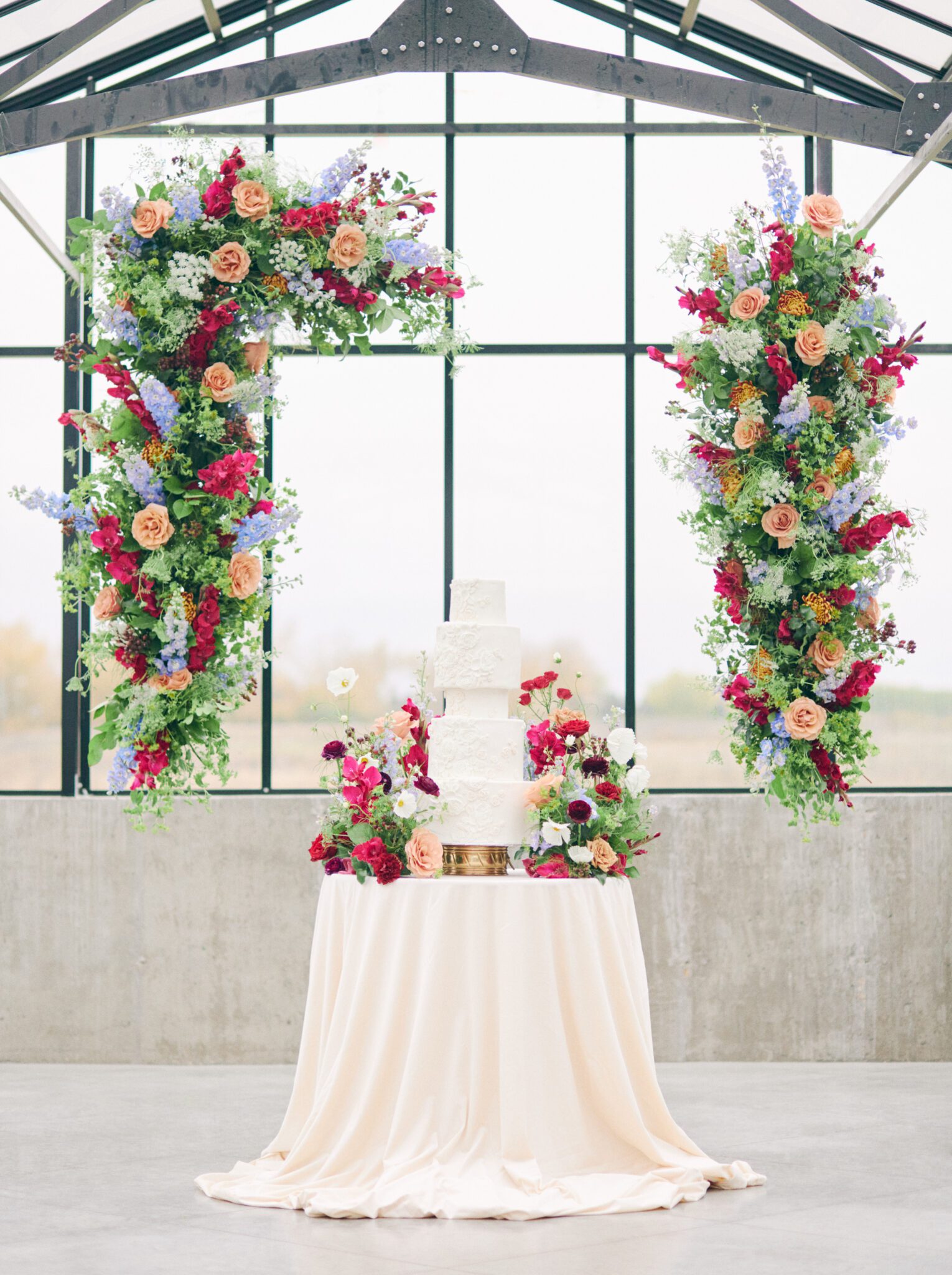 Elegant five-tiered wedding cake with iced floral details, surrounded by dramatic and colourful florals in pink, peach, orange, and violet at The Station’s solarium in Saskatchewan.
