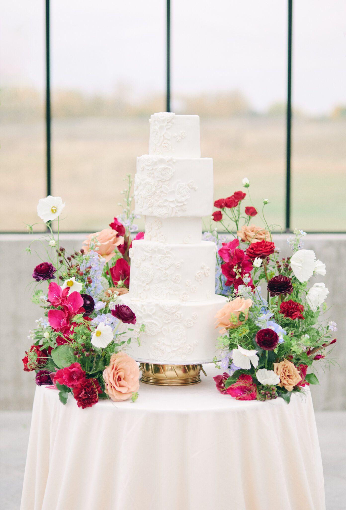 Elegant five-tiered wedding cake with iced floral details, surrounded by dramatic and colourful florals in pink, peach, orange, and violet at The Station’s solarium in Saskatchewan.
