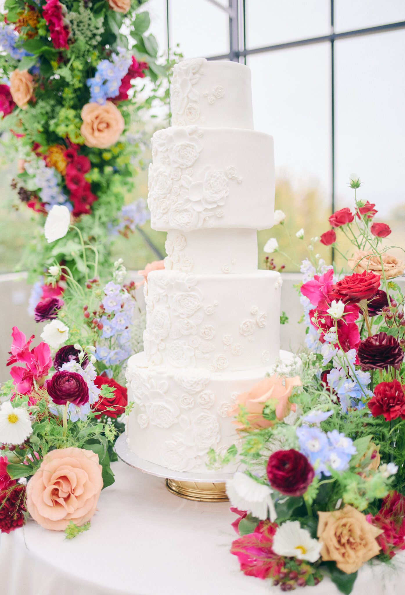 Elegant five-tiered wedding cake with iced floral details, surrounded by dramatic and colourful florals in pink, peach, orange, and violet at The Station’s solarium in Saskatchewan.
