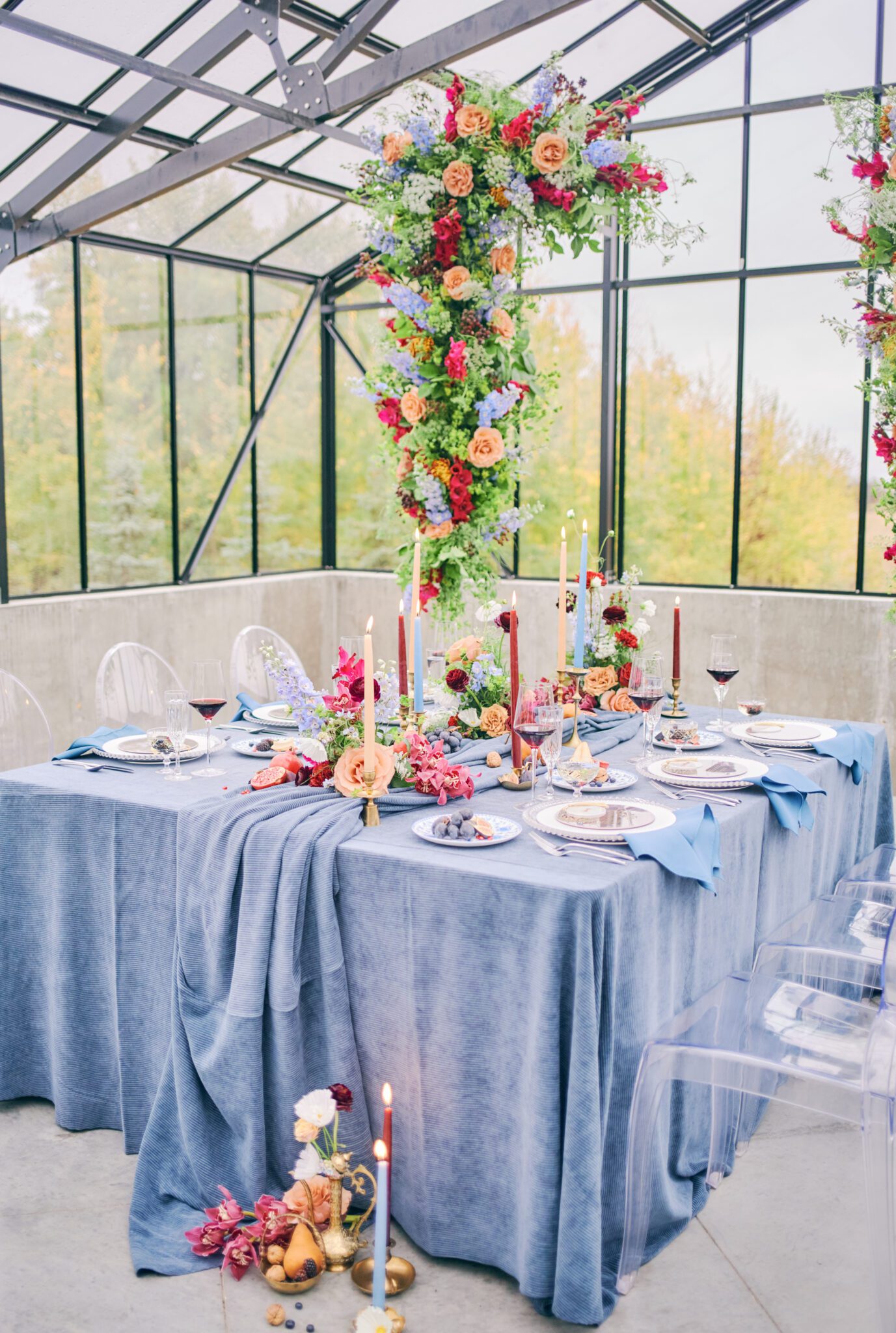 Wedding reception tablescape featuring vibrant flowers in pink, peach, violet, and red, blue corduroy linens, colorful candles with hints of gold, and fruit. Floating floral arch framing the intimate reception space.