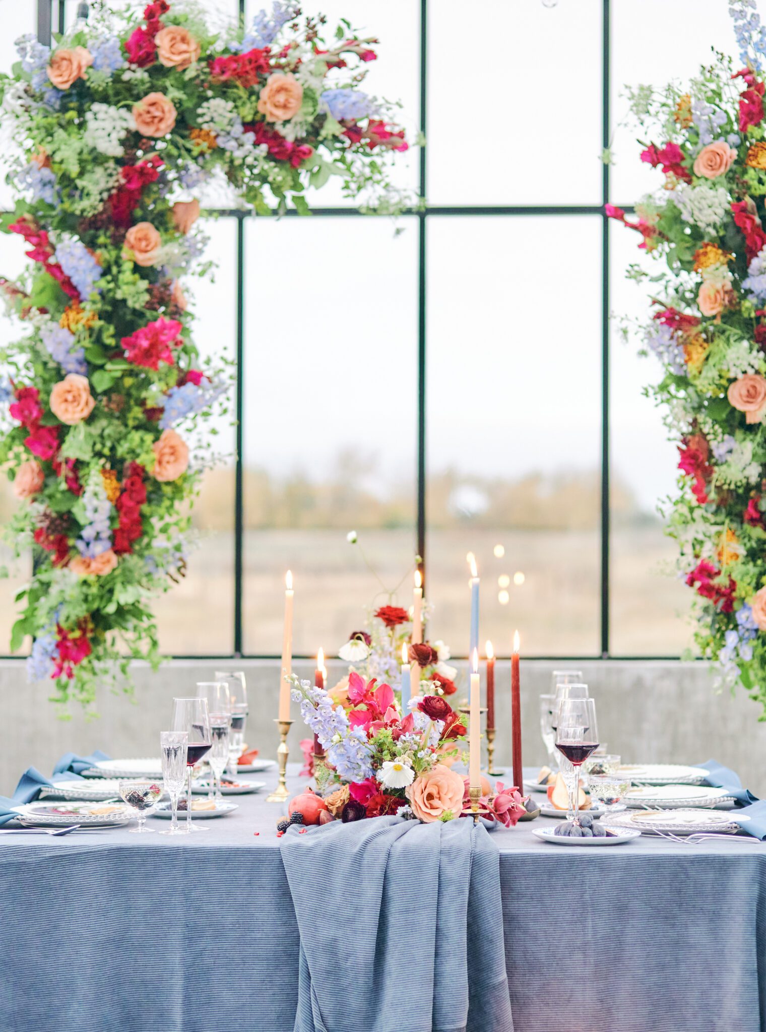 Wedding reception tablescape featuring vibrant flowers in pink, peach, violet, and red, blue corduroy linens, colourful candles with hints of gold, and fruit. Floating floral arch framing the intimate reception space.