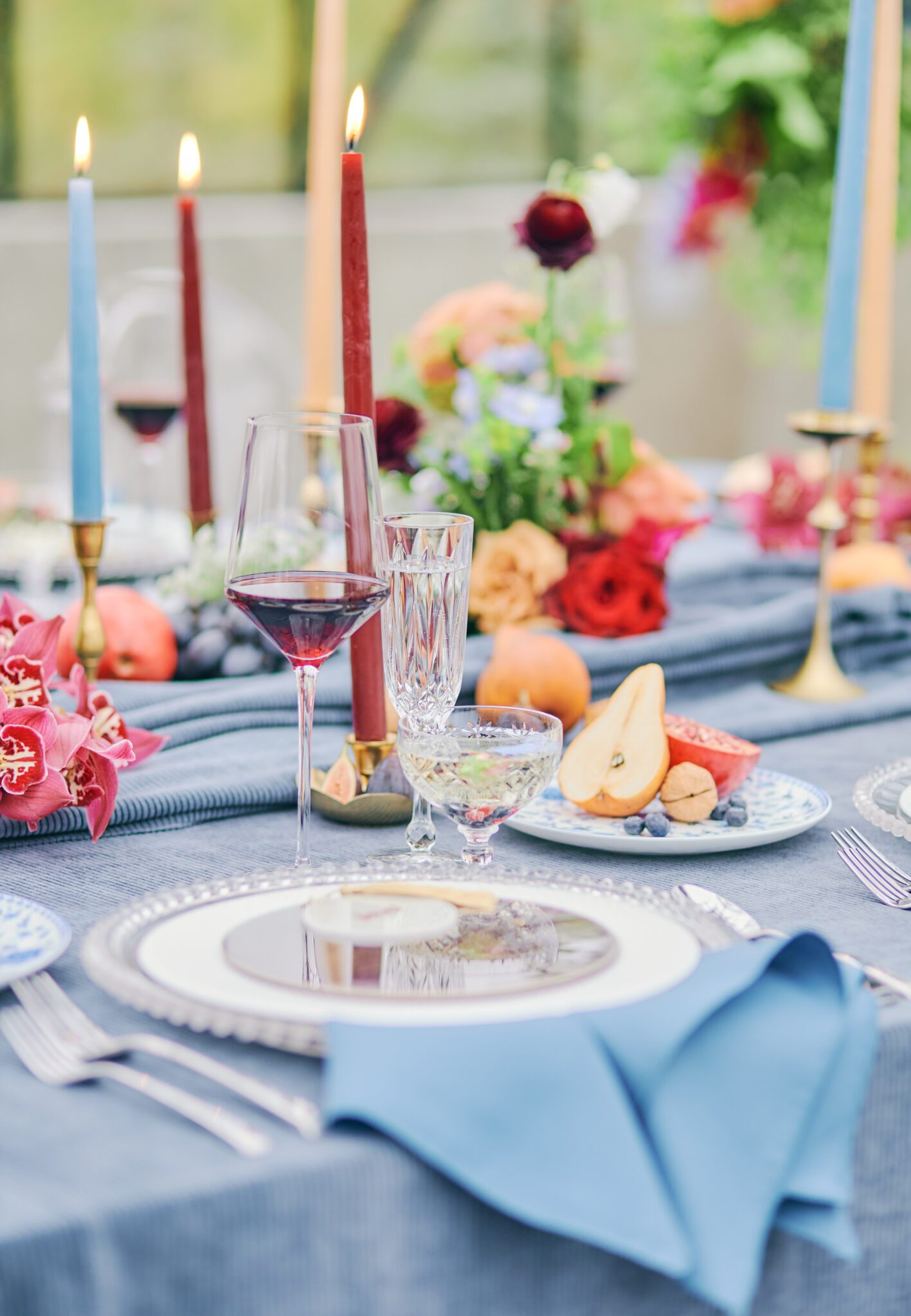 Wedding reception tablescape featuring vibrant flowers in pink, peach, violet, and red, blue corduroy linens, colourful candles with hints of gold, and fruit.