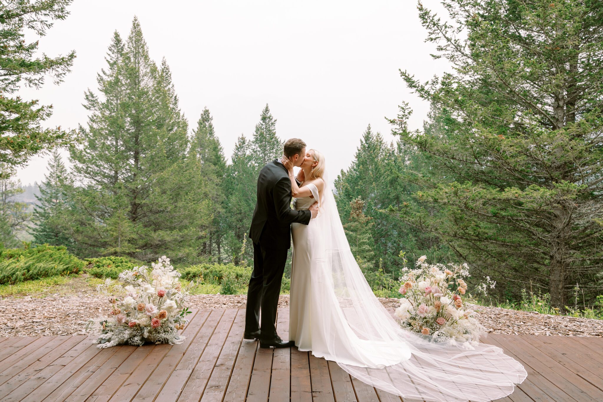 Bride and groom kissing during wedding ceremony at Stewart Creek Golf Course in Canmore, Alberta. White, peach and pink florals. Mountain wedding ceremony. 