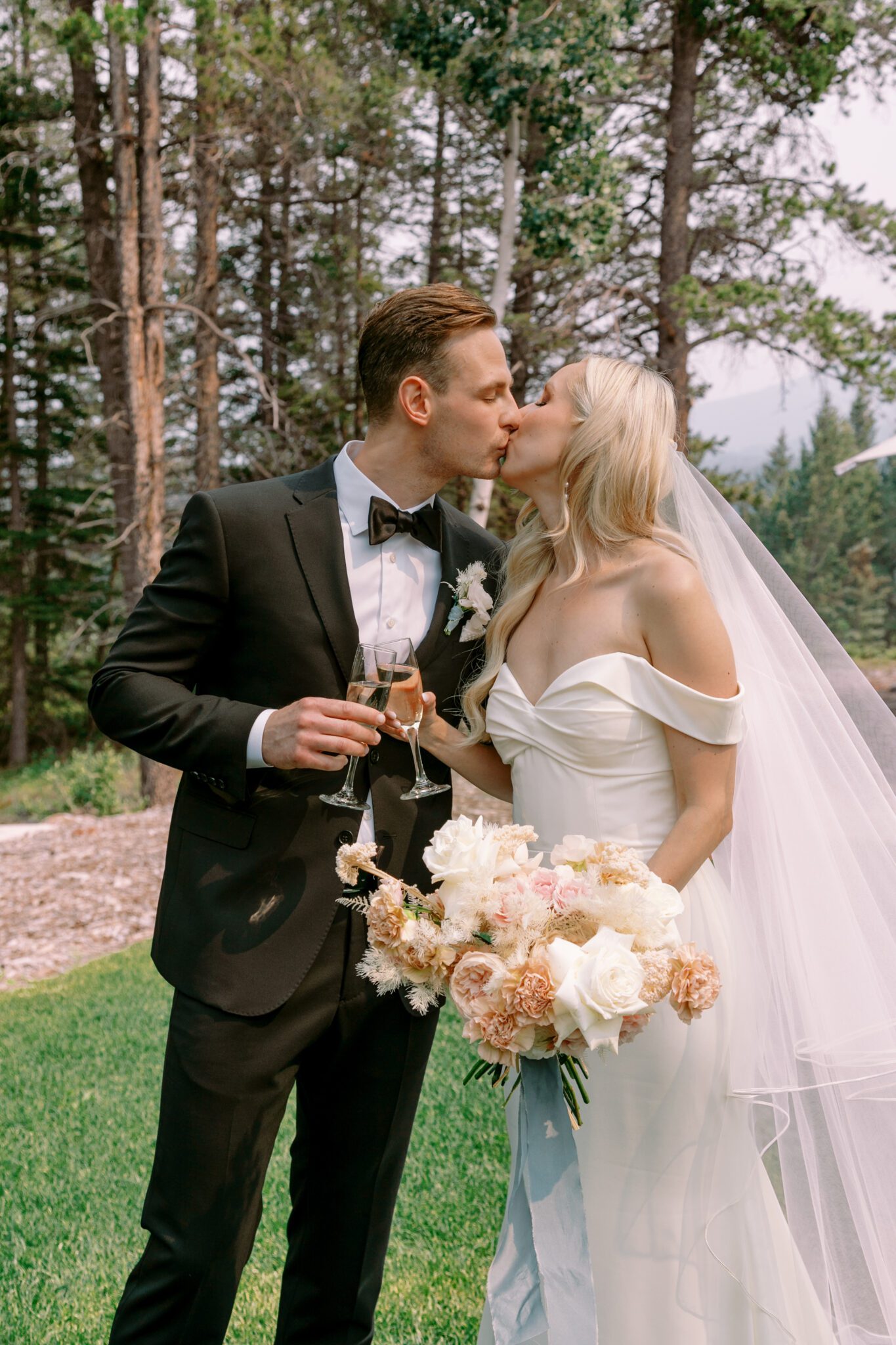 Bride and groom kissing with prosecco during Stewart Creek Golf Course Wedding in Canmore, Alberta. Pink, peach and white floral bouquet tied with teal silk ribbon. Bride wearing sleek, form-fitting bridal gown with long veil.