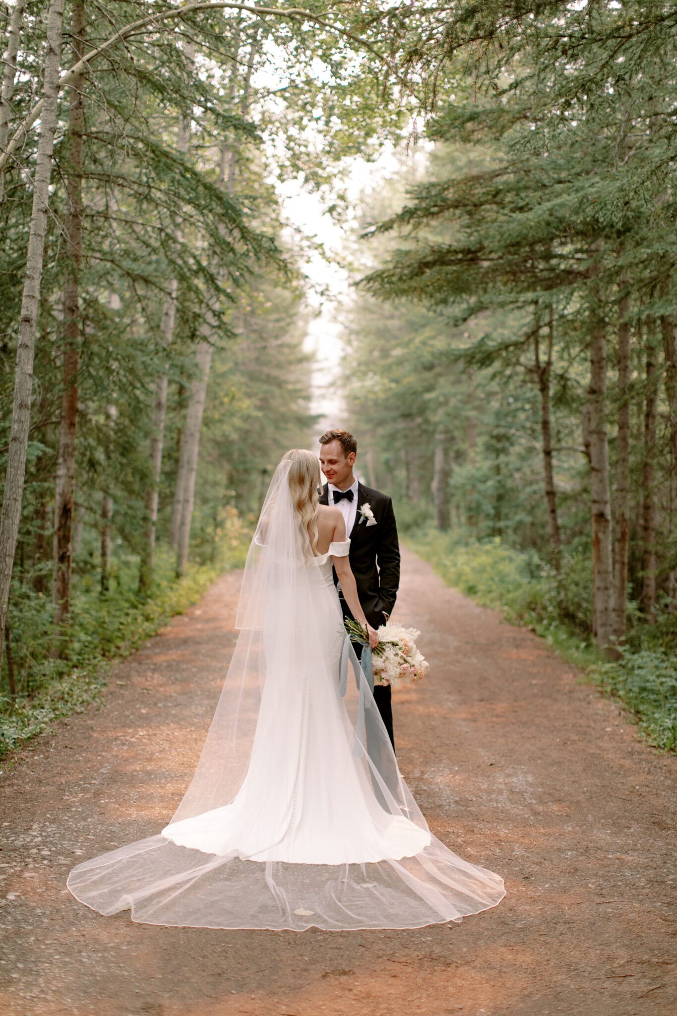 Bride and groom embracing during Steward Creek Golf Course wedding in Canmore, Alberta. Spring wedding inspiration. Peach and white floral bouquet tied with teal silk ribbon. The bride wearing a sleek, form-fitting bridal gown with a long cathedral veil.