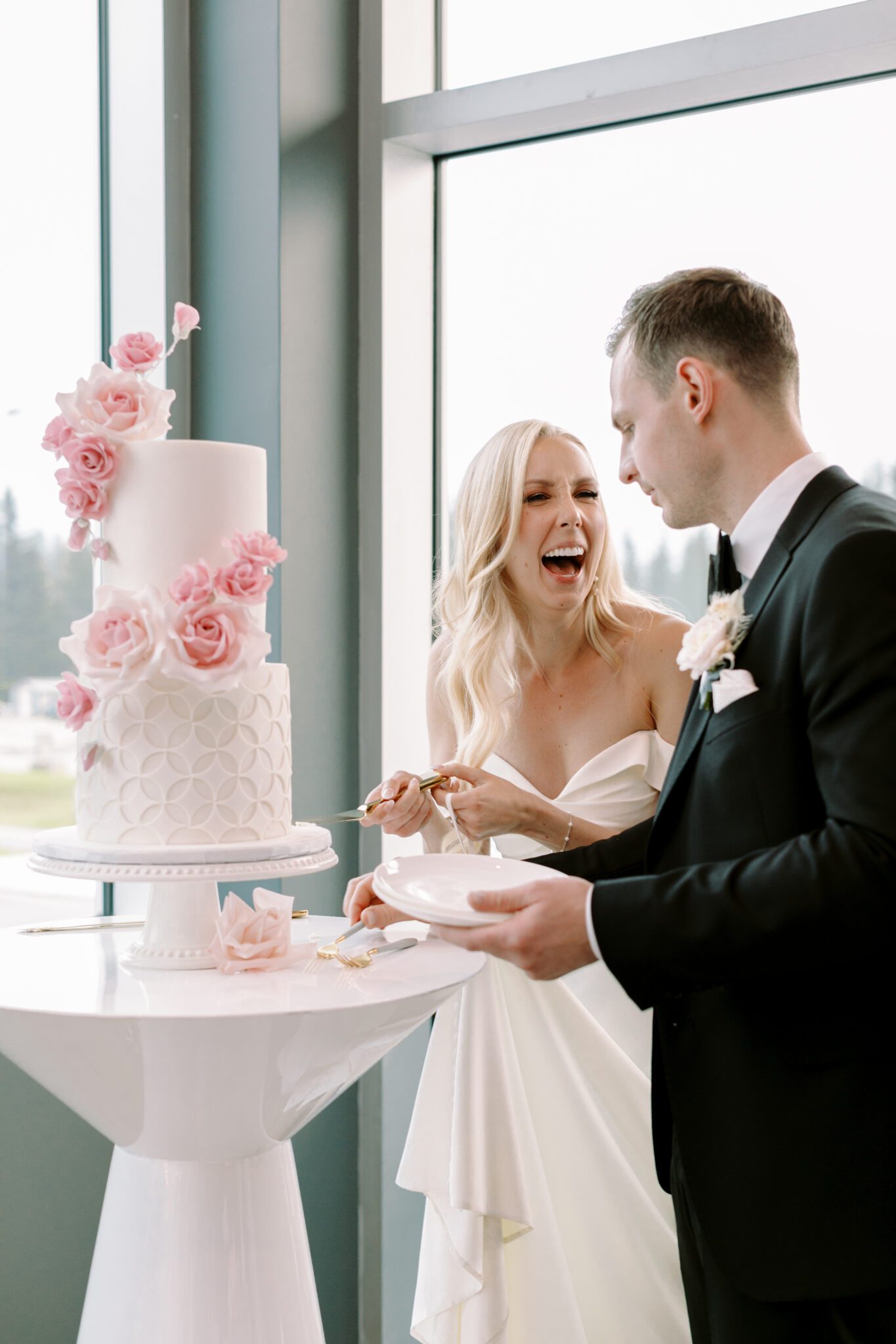 Bride and groom cutting a beautiful 2-tiered white wedding cake with pink sugar roses created by Yvonne's Delightful Cakes.