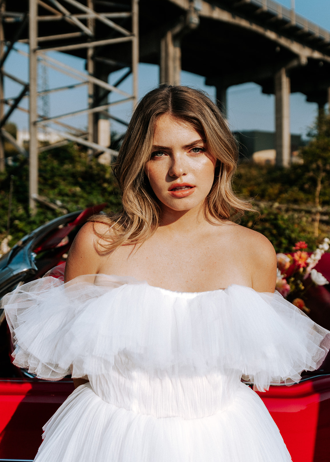 Carefree bad-ass bride wearing flirty Zephyr dress by local designer Ouma Bridal, standing in front of vintage red muscle car surrounded by tangerine and white summer blooms. 